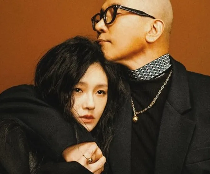 Stylish couple in black coats poses against a warm brown background in a moody fashion portrait.