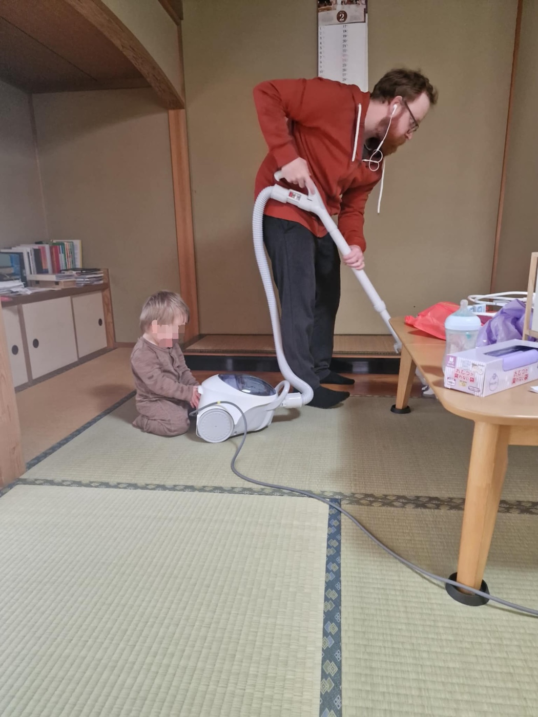 Bildet viser en mann som støvsuger, mens et barn sitter på gulvet bak