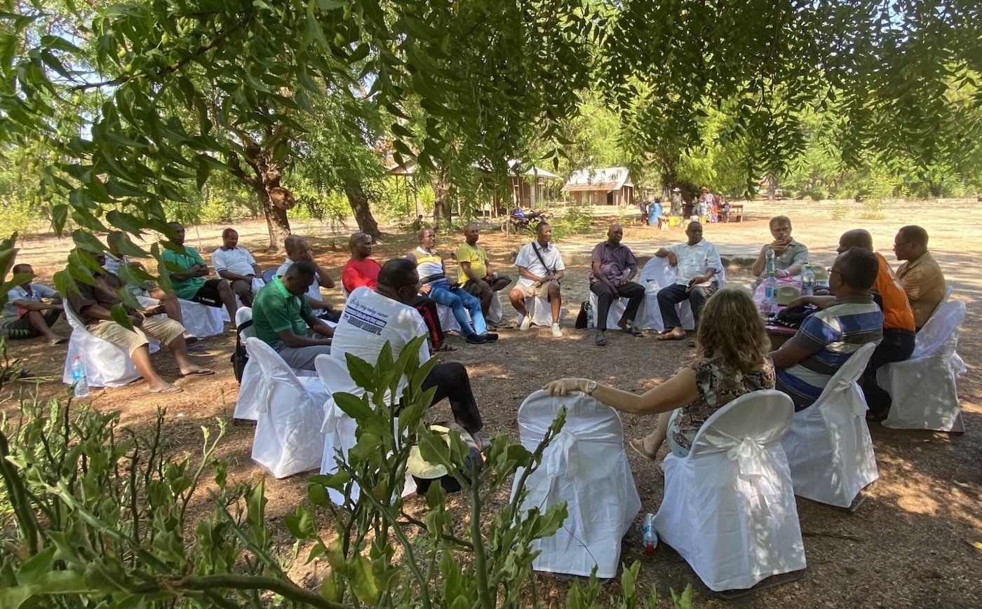 Bildet viser en forsamling mennesker som sitter på stoler i ring utendørs, under grønne, frodige trær