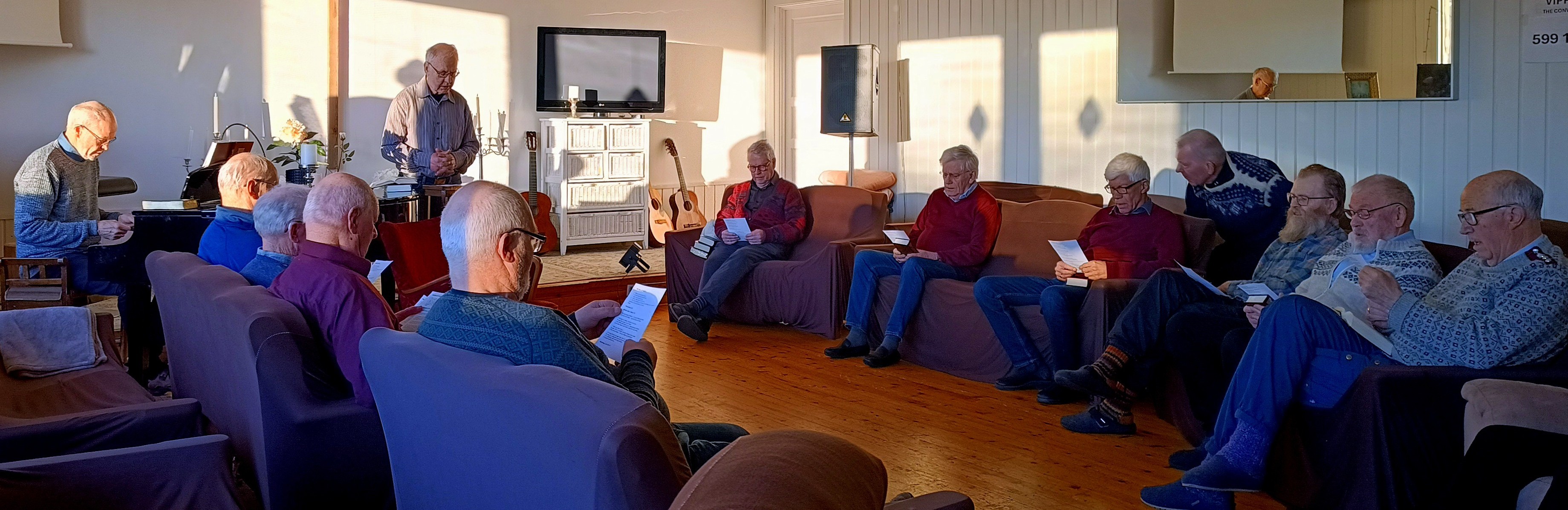 Bildet viser en gruppe menn som sitter i stoler og sofaer og synger