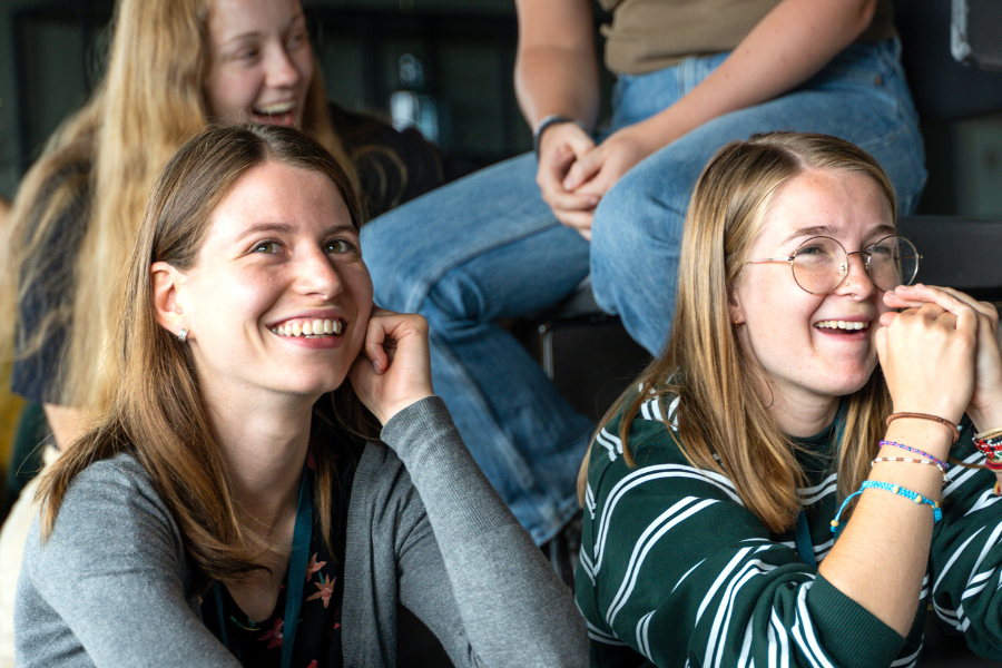 Bildet viser to personer som smiler mens de sitter og følger med på noe som skjer foran dem