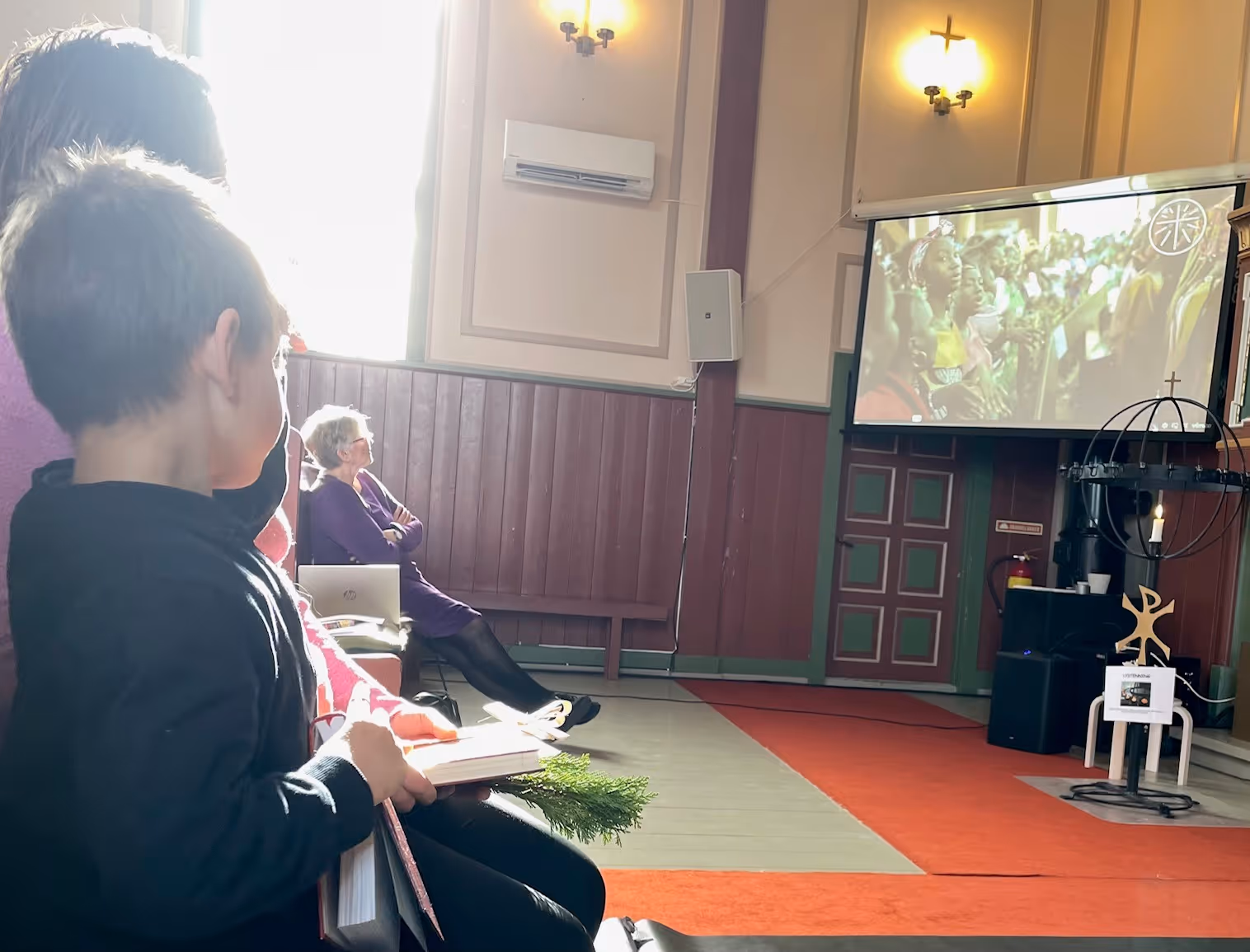 Bildet viser personer som sitter og ser en video på en TV-skjerm i et kirkerom