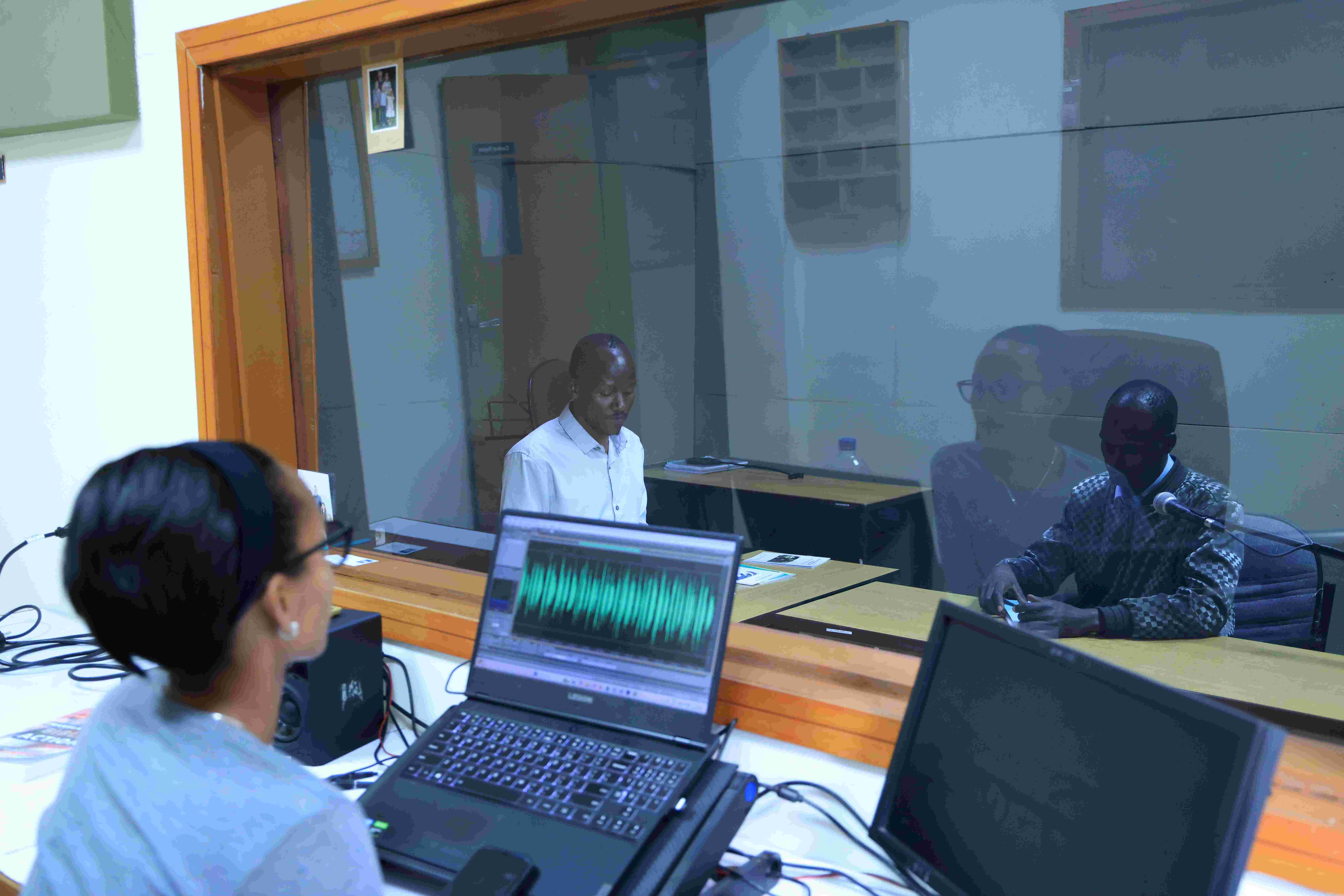 Bildet viser to personer inne i et radiostudio, mens en rodusent overvåker lydbildet fra en PC i et tilstøtende rom