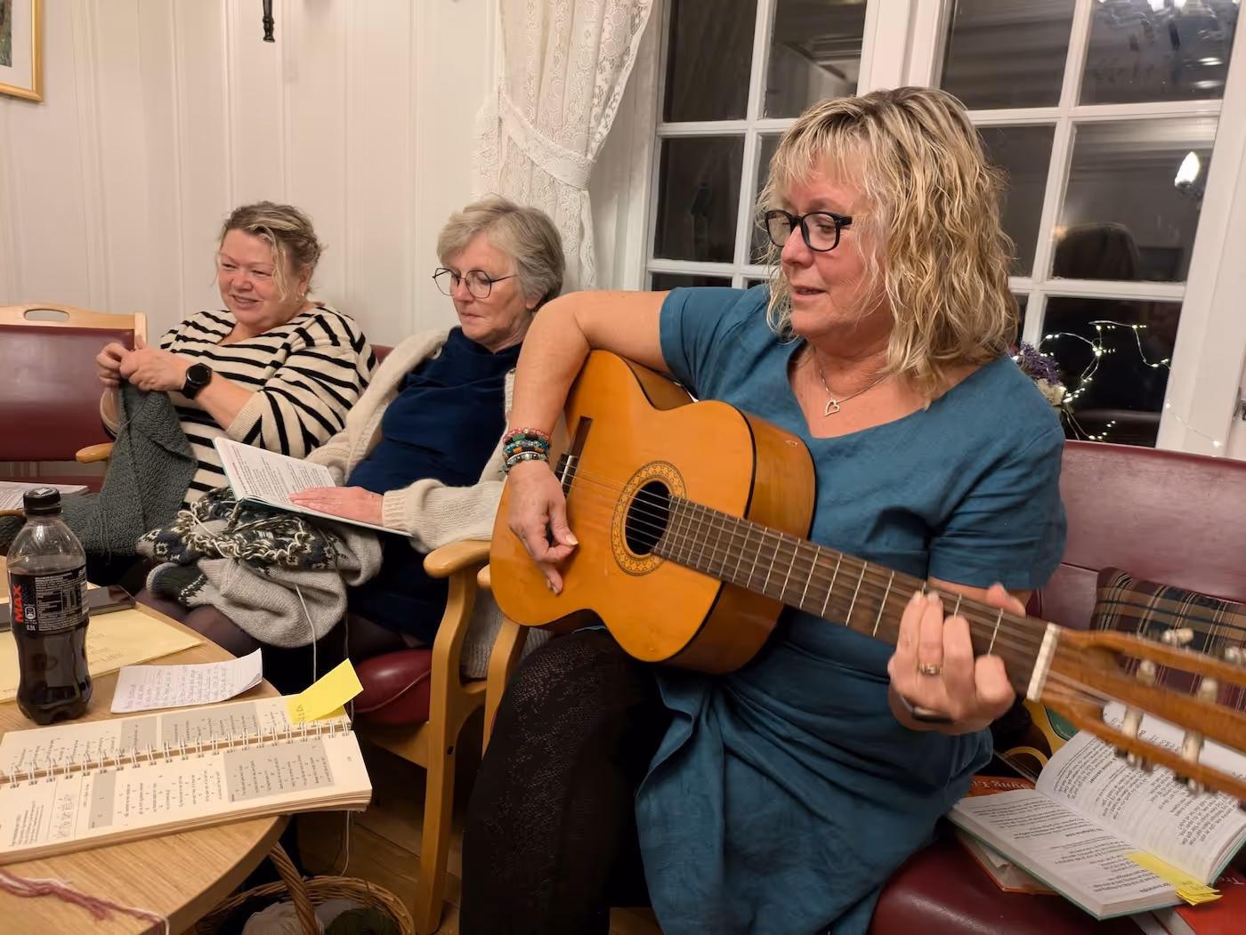 Bildet viser tre damer som sitter i en sofa. amen til venstre strikker, damen i midten leser eller syger entekst fra et ark, og damen til høyre spiller på en gitar