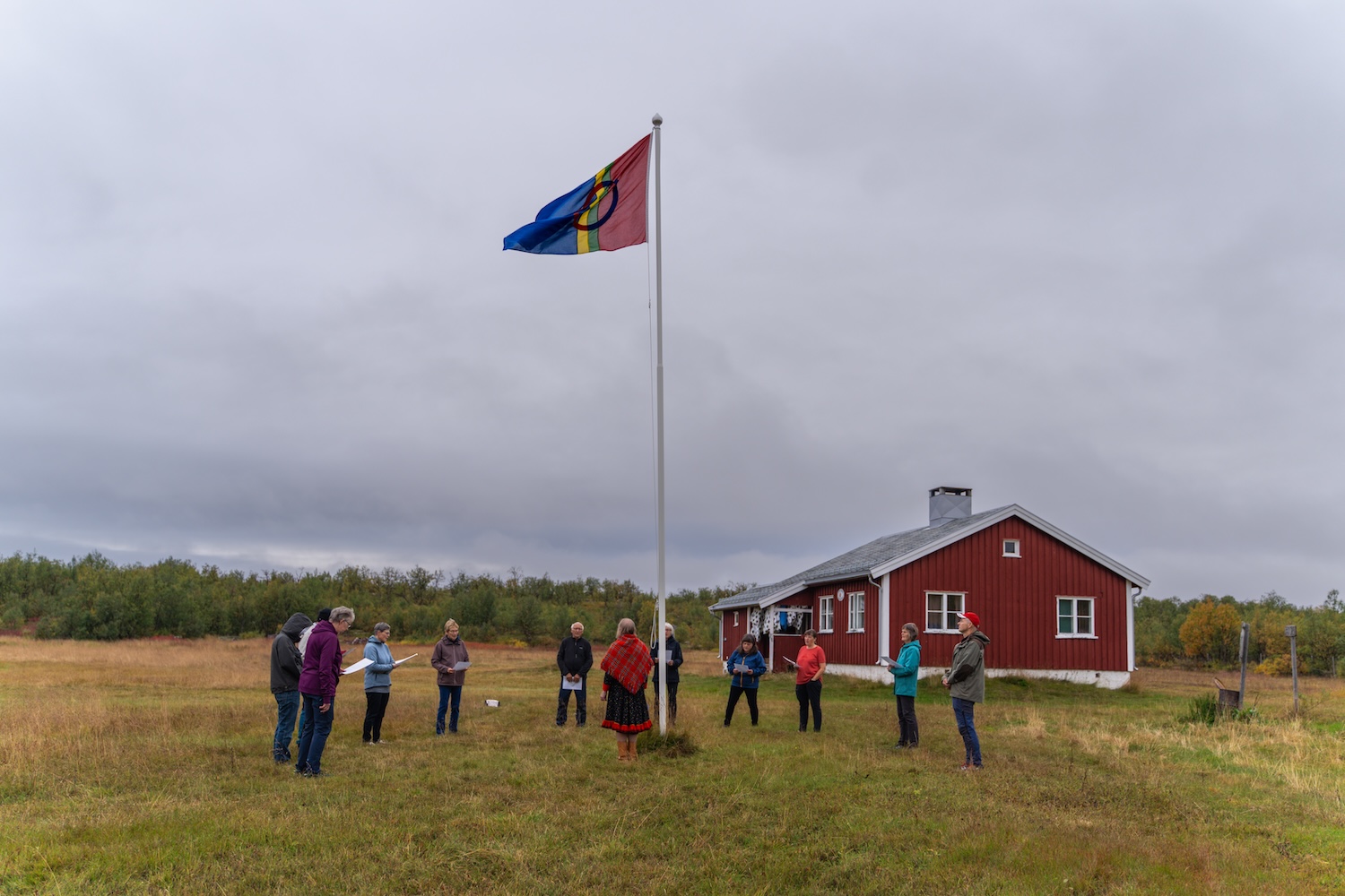 Bildet viser 12 personer stående rundt ei flaggstang med det samiske flagget heist til topps