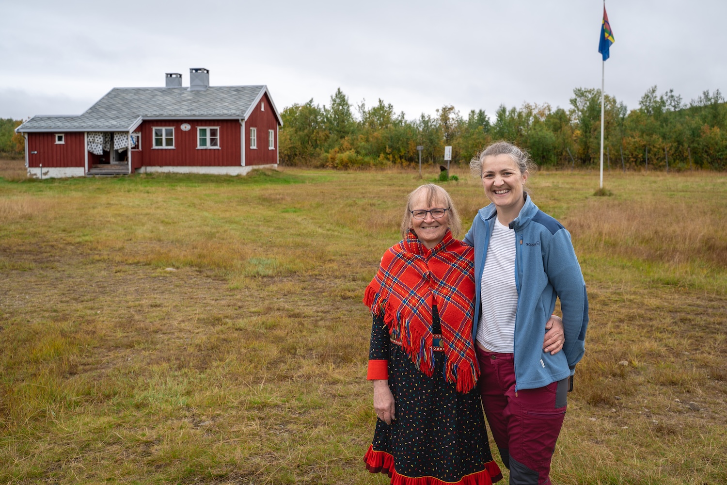Bildet viser to personer stående utendørs foran et rødmalt hus til venstre, og ei flaggstang med det samiske flagget til høyre