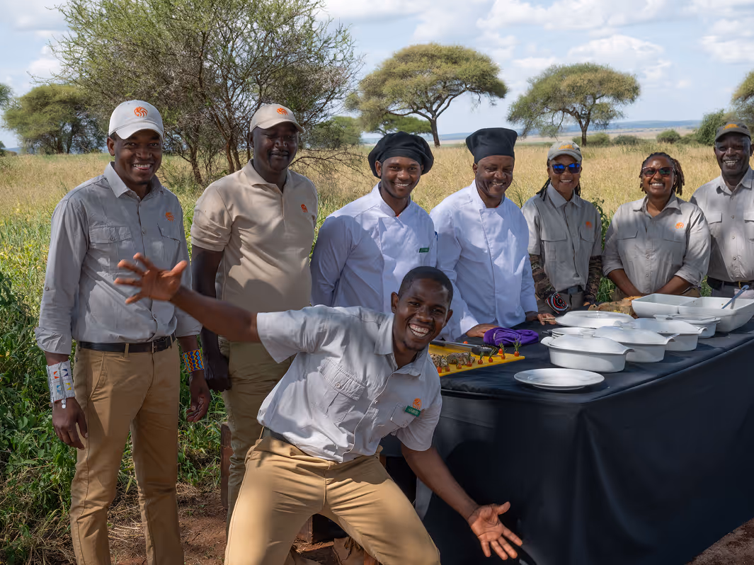 African staff team smiling outdoors with diverse dishes on a colorful buffet display.