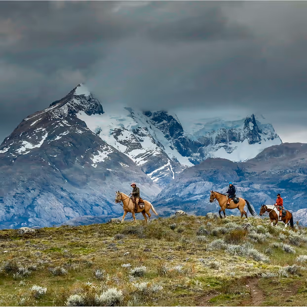 Explorers on horseback trek through Patagonia's breathtaking mountains with snow-capped peaks and cloudy skies.