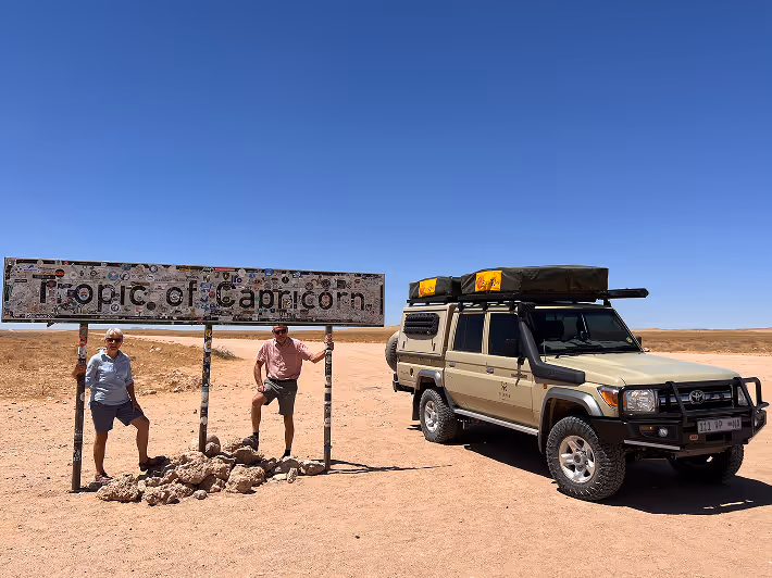 Men posing boldly at Tropic of Capricorn sign with rugged off-road vehicle adventure.