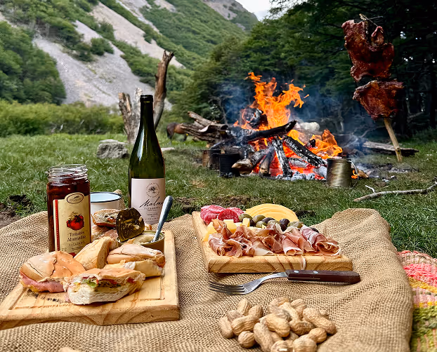 Cozy outdoor picnic setup near campfire featuring wine, meats, cheeses, and snacks gathered.
