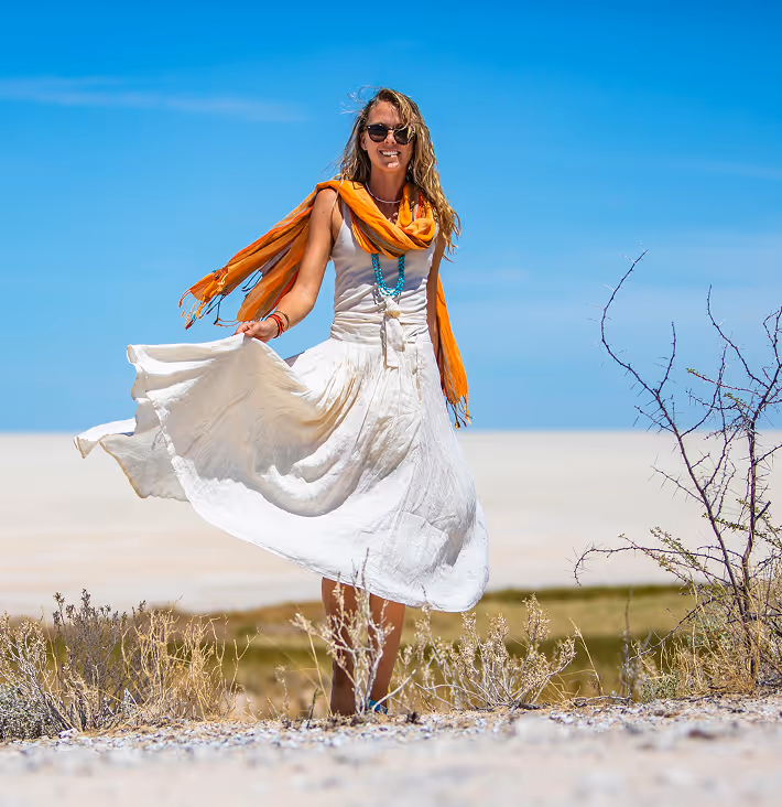 Woman in white dress twirling with orange scarf in desert landscape against blue sky backdrop.