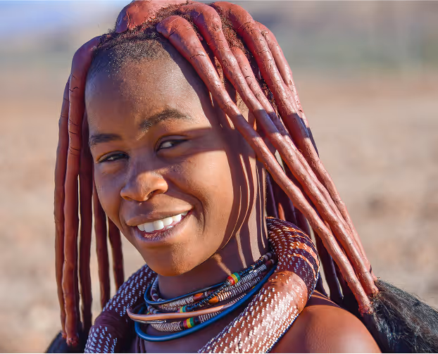 African woman with vibrant red dreadlocks and colorful beaded jewelry in a natural outdoor setting.