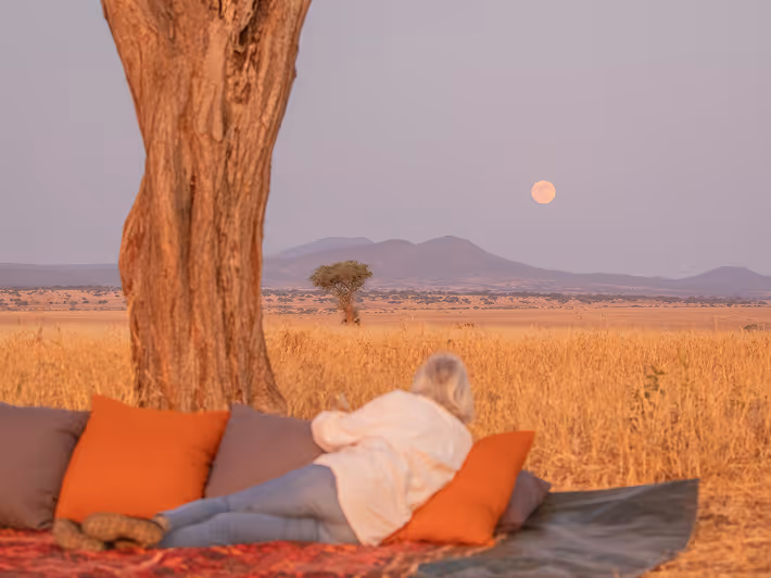 Relaxed woman soaks in the serene African savannah sunset under a majestic full moon.