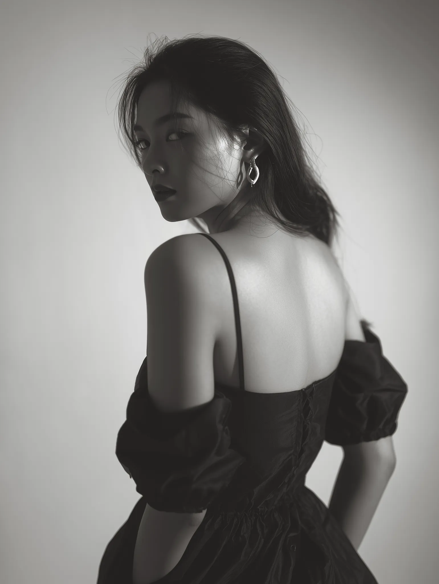 Black and white portrait of a woman in an off-shoulder black dress, glancing over her shoulder with a confident and enigmatic expression, softly lit against a neutral background.