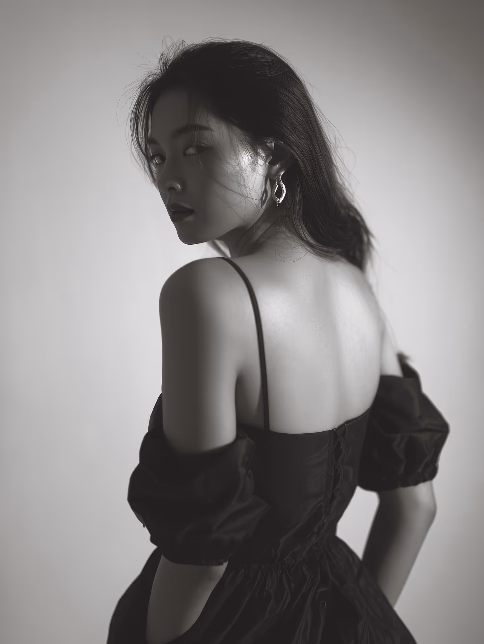 Black and white portrait of a woman in an off-shoulder black dress, glancing over her shoulder with a confident and enigmatic expression, softly lit against a neutral background.