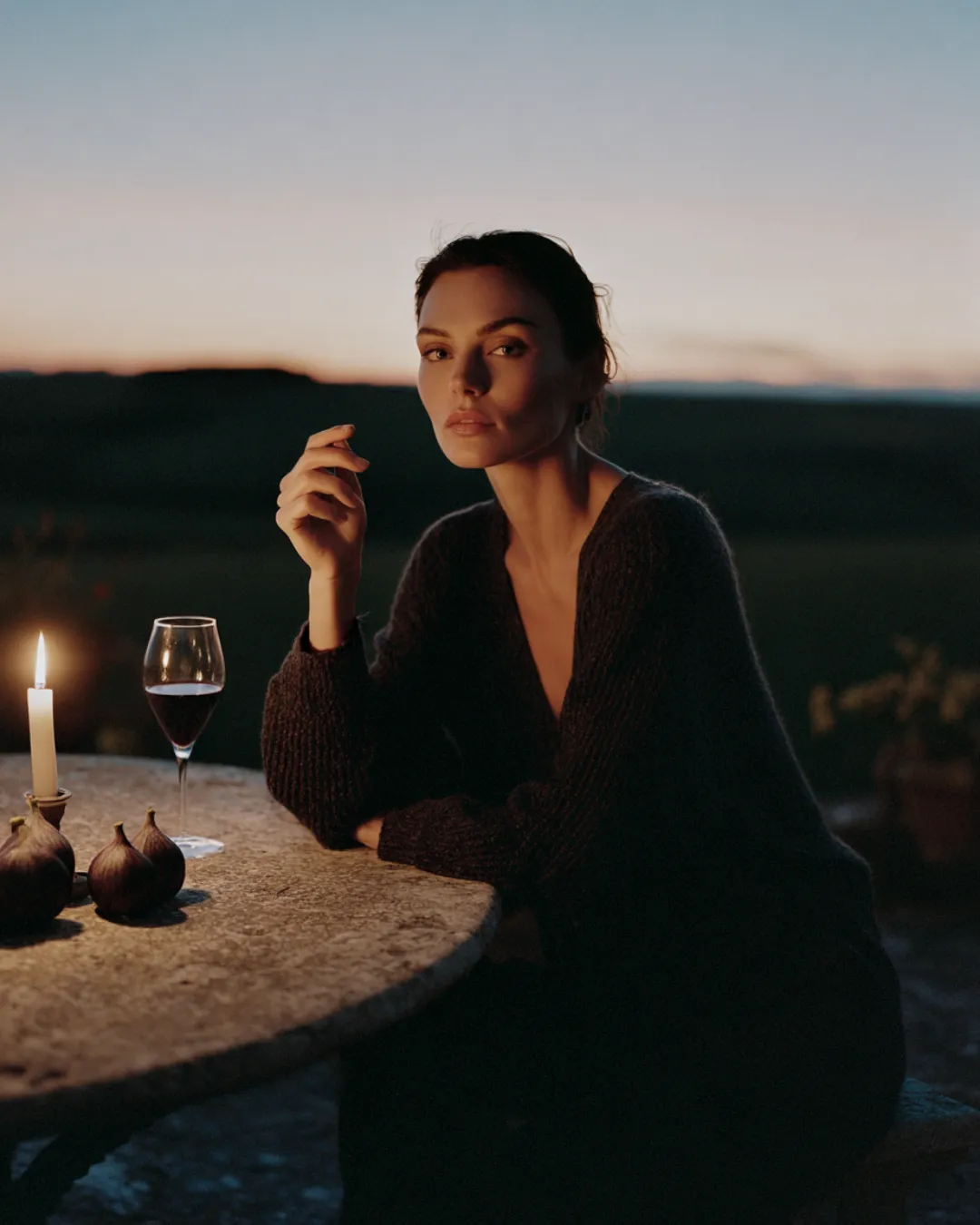 A woman in a dark sweater sits at a stone table at dusk, a candle and a glass of red wine beside her — evoking evening stillness and reflective conversation.