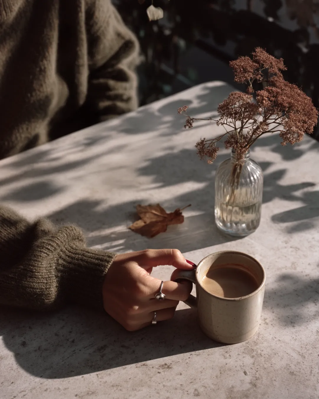 A hand holding a ceramic cup of coffee rests on a marble table next to a small glass vase with dried flowers — a quiet autumnal moment in soft morning light.