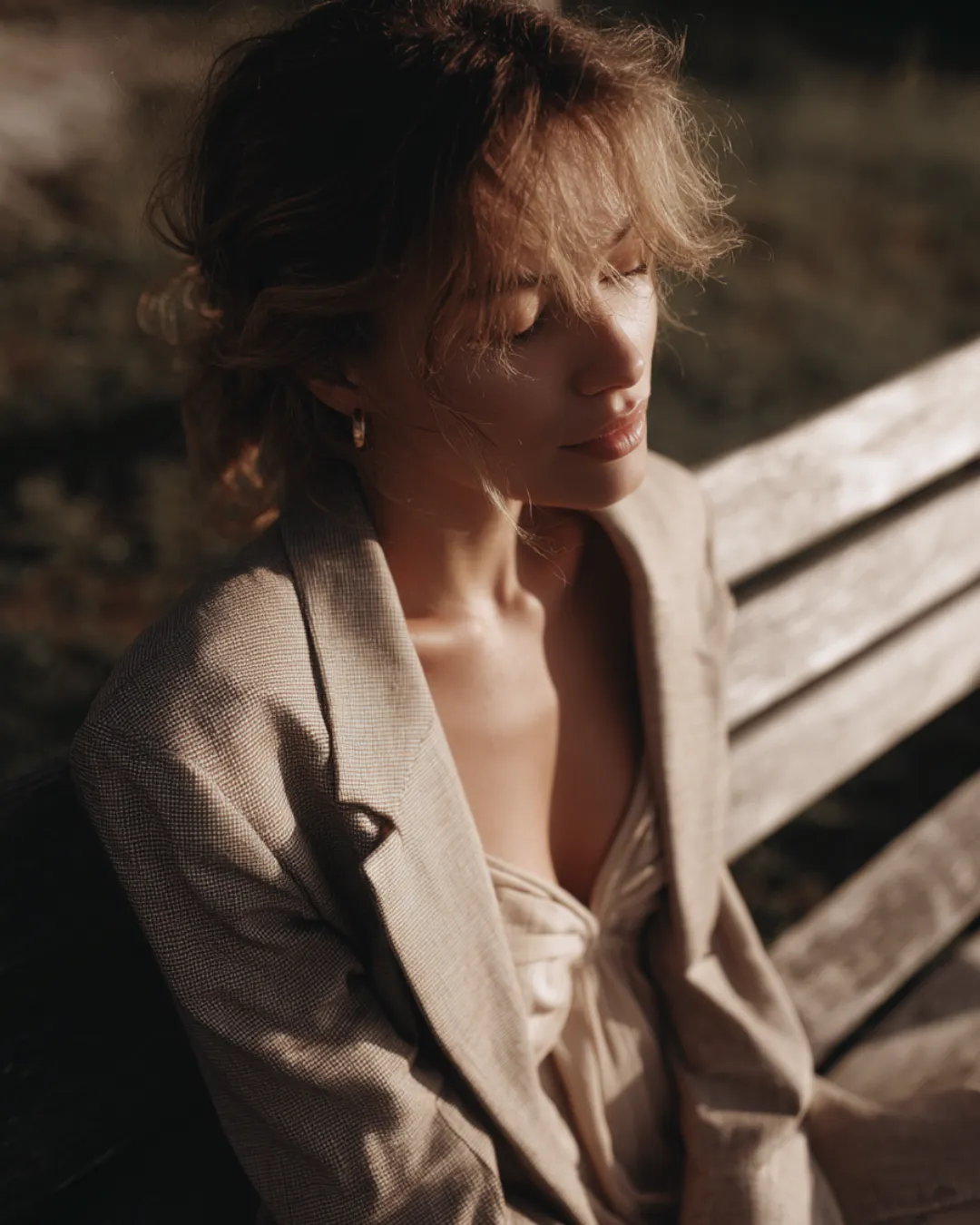A woman sitting on a wooden bench with her eyes closed, dressed in a neutral-toned blazer and softly lit by warm sunlight — capturing a moment of calm introspection.