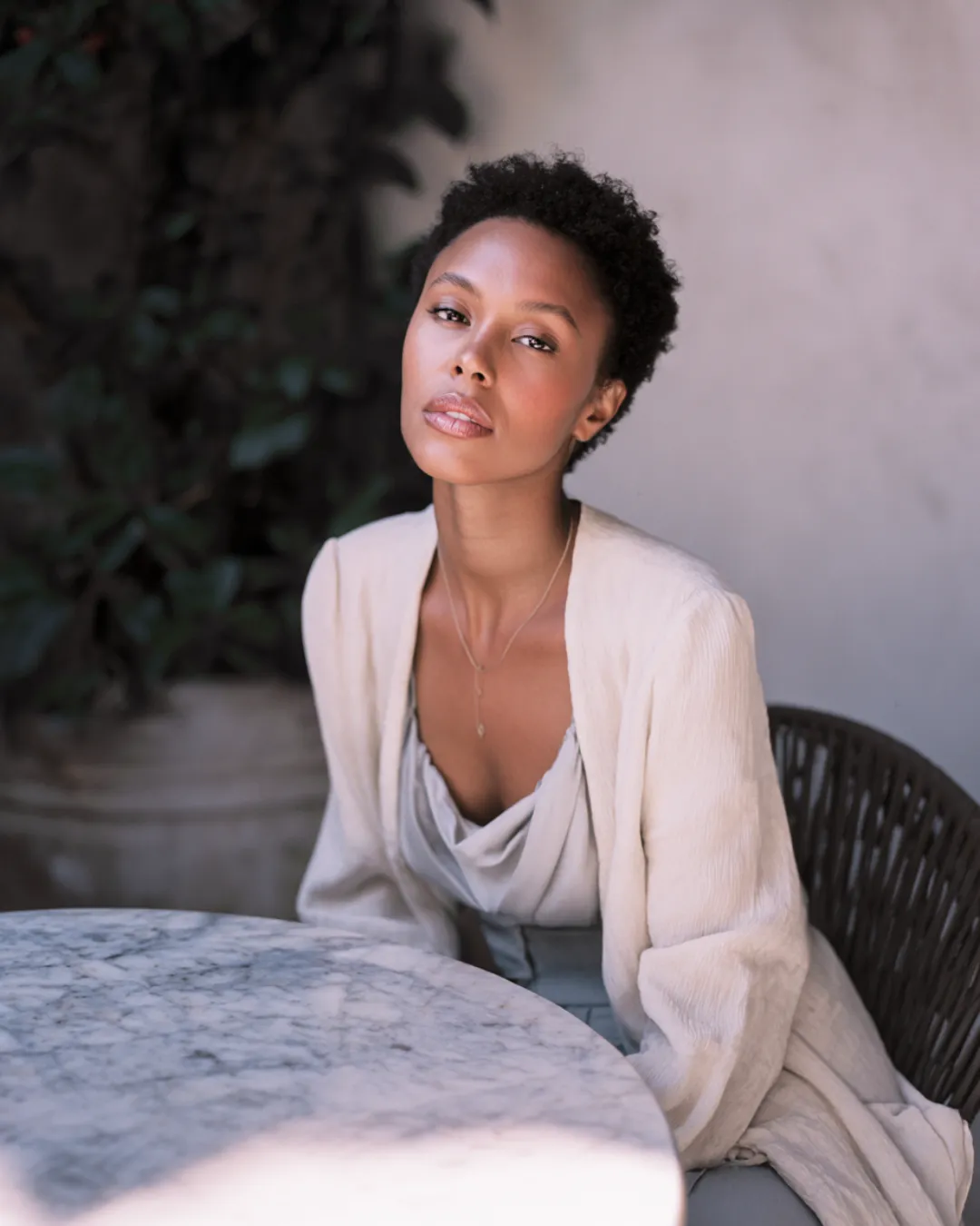 A woman seated at a round marble table, wearing a light, flowy outfit and gazing calmly at the camera — an elegant portrait set against a soft, minimalist backdrop.
