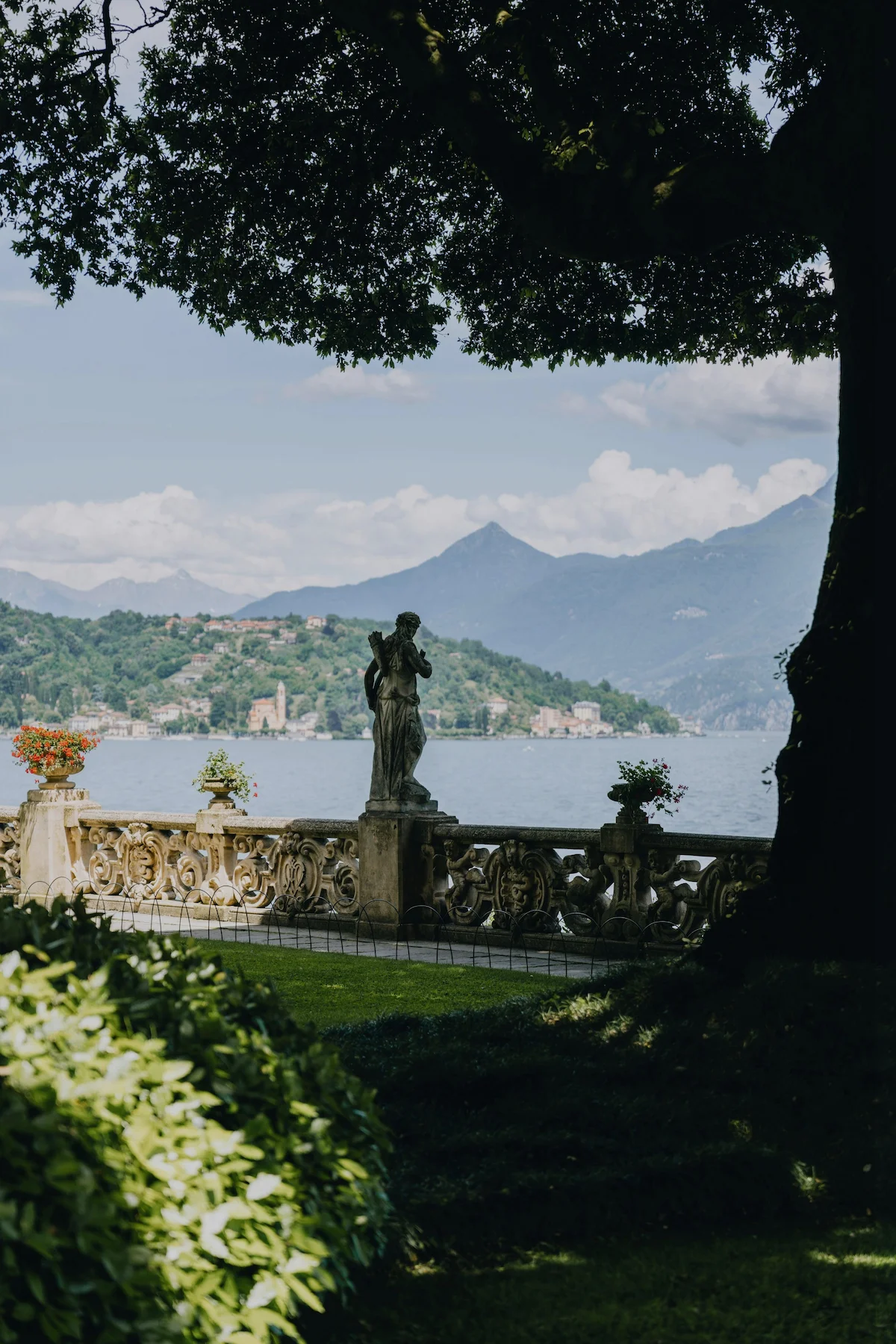 Stone statue on ornate balustrade overlooking a lake with mountains and a village in the background, framed by large tree branches and greenery.