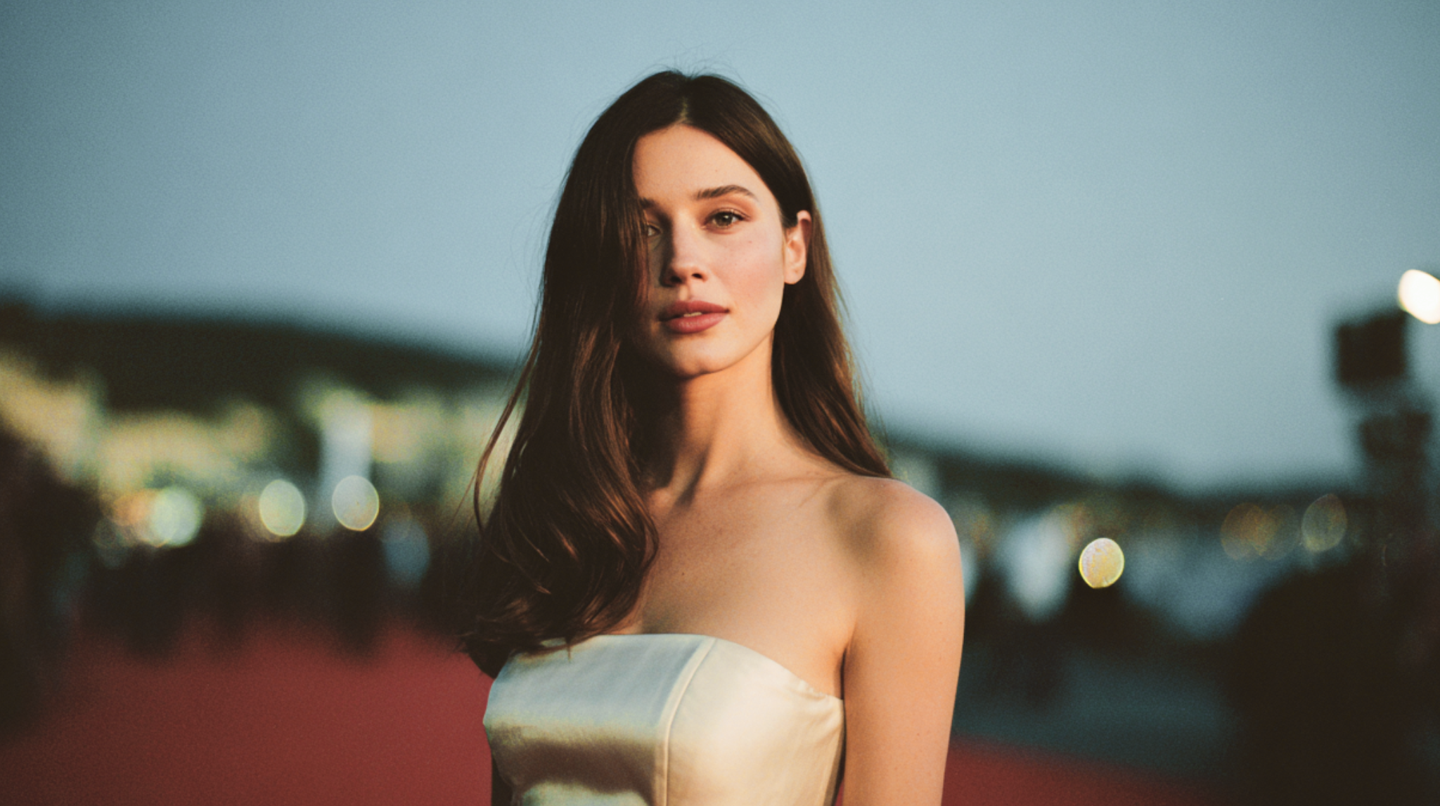 A woman on a red carpet at dusk, wearing a minimalist silk gown, radiating understated elegance and presence.