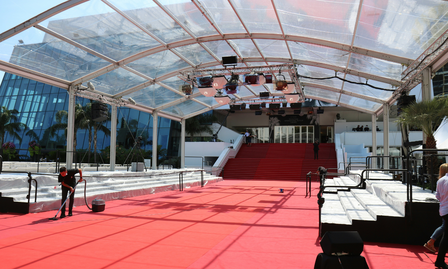 Behind-the-scenes view of the red carpet setup at the Cannes Film Festival 2025, showing a transparent canopy structure, red stairs leading to the entrance, and staff preparing the area under bright daylight.