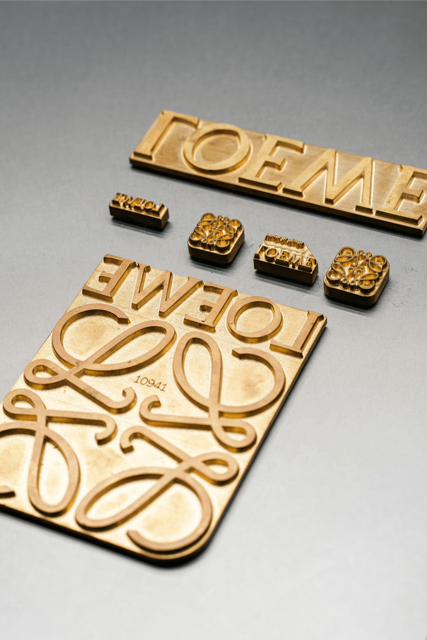 A set of gold metal stamps featuring the Loewe logo and typography arranged on a smooth grey surface. The refined craftsmanship reflects the brand’s dedication to detail and heritage in luxury branding.