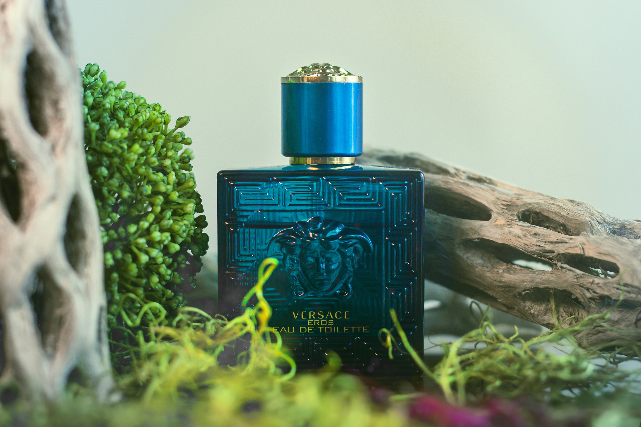 A luxurious deep blue bottle of "Versace Eros Eau de Toilette" placed among lush green moss and weathered driftwood. The natural surroundings highlight the elegance and sensuality of the perfume's golden details.