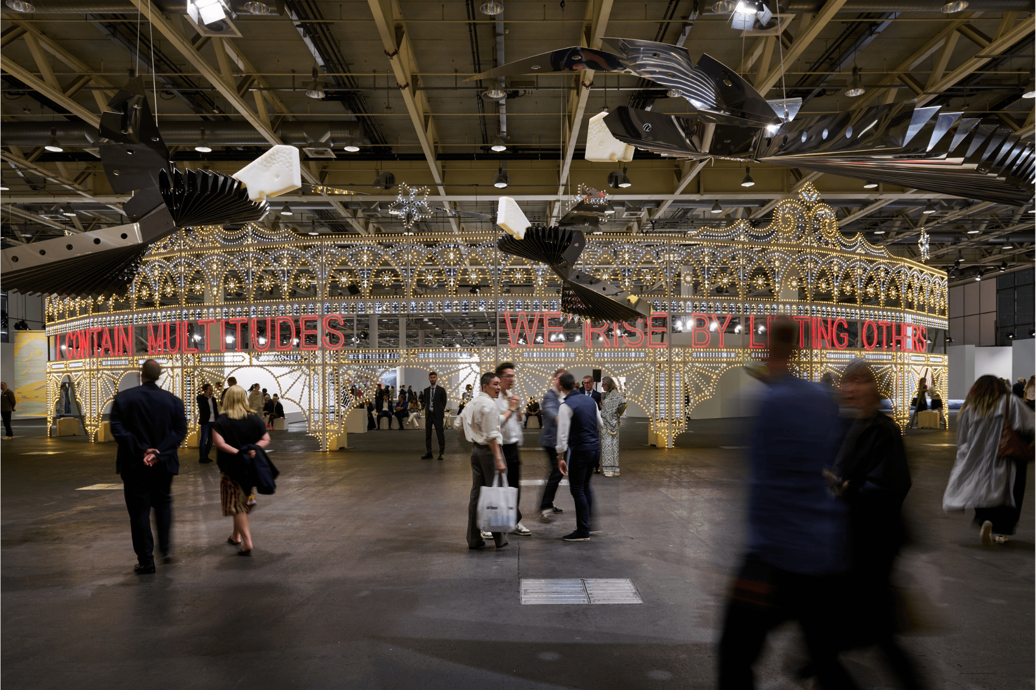 Large illuminated installation with the phrase “I CONTAIN MULTITUDES – WE RISE BY LIFTING OTHERS” at Art Basel 2025. Visitors walk through the exhibition space under an industrial-style ceiling with overhead lighting.