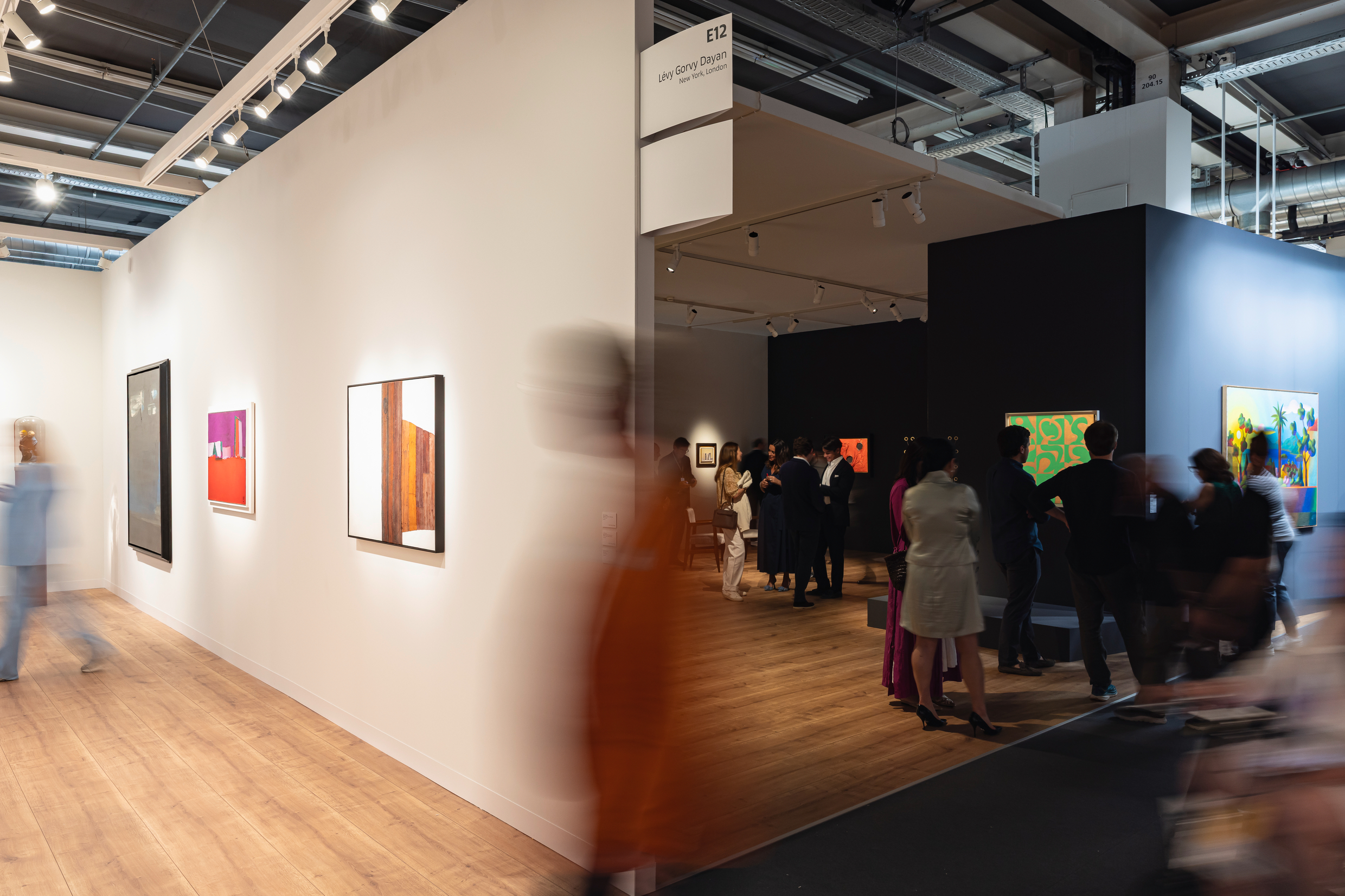 Art fair visitors walk and interact inside a brightly lit gallery booth at Art Basel 2025. The space features modern paintings, wood flooring, and a mix of white and dark exhibition walls, with people blurred in motion.