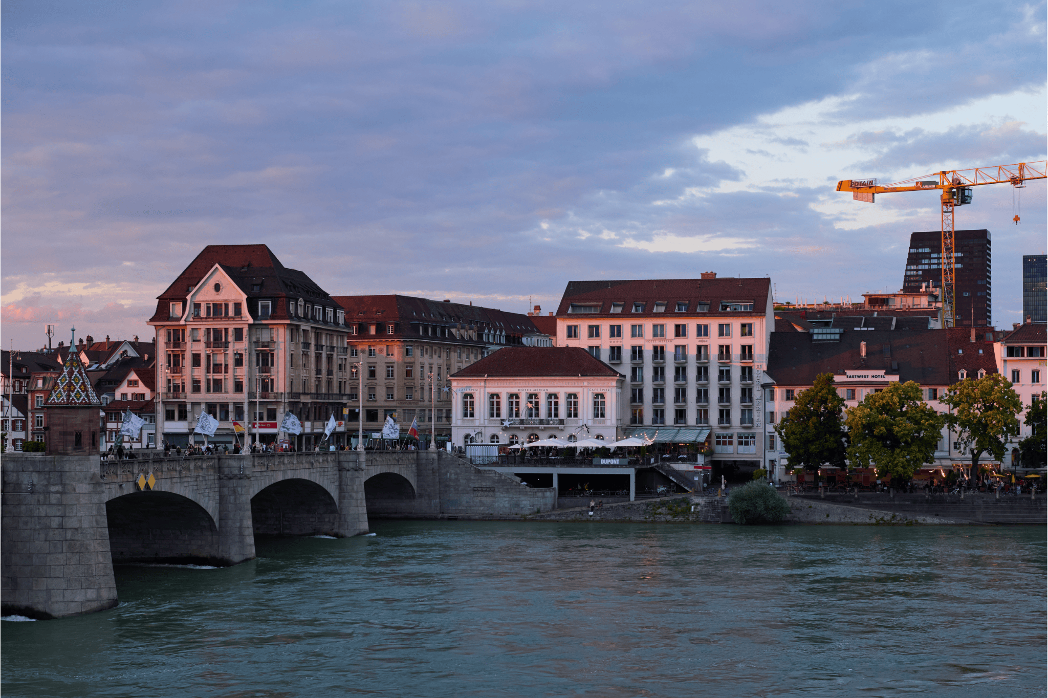 A scenic riverside view of Basel at dusk during Art Basel 2025. The Mittlere Brücke bridge is lined with flags and people, historic buildings glow in soft evening light, and a construction crane rises in the background.