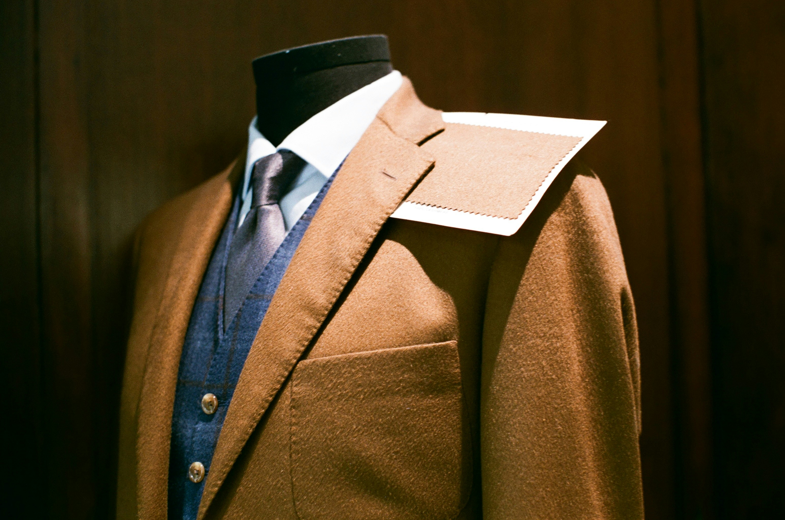 Elegant brown tailored suit with blue waistcoat and tie on a mannequin, featuring a fabric swatch on the shoulder, showcased at Pitti Uomo 2025 fashion fair