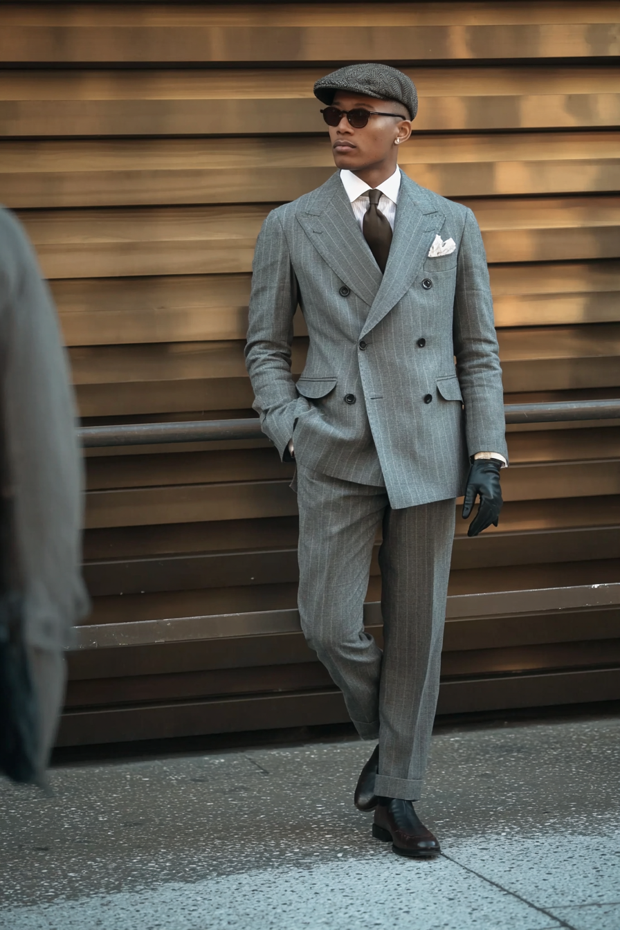 Stylish man in a tailored grey double-breasted pinstripe suit with black gloves, flat cap, and sunglasses posing against a metallic background at Pitti Uomo 2025