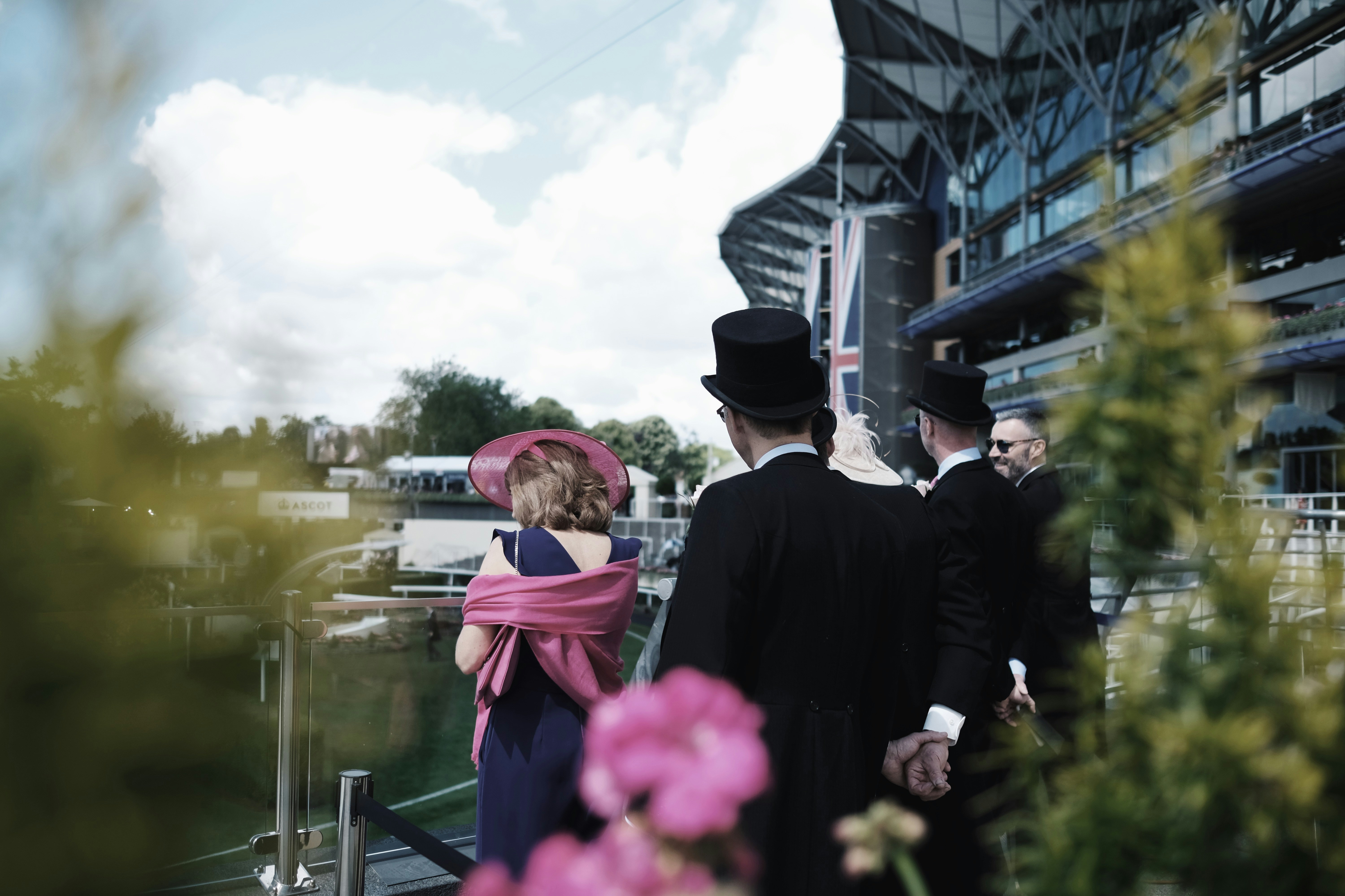 Well-dressed spectators in formal attire, including top hats and a pink fascinator, overlooking the racetrack at Royal Ascot 2025 from a grandstand terrace, with the Union Jack and modern architecture in the background.