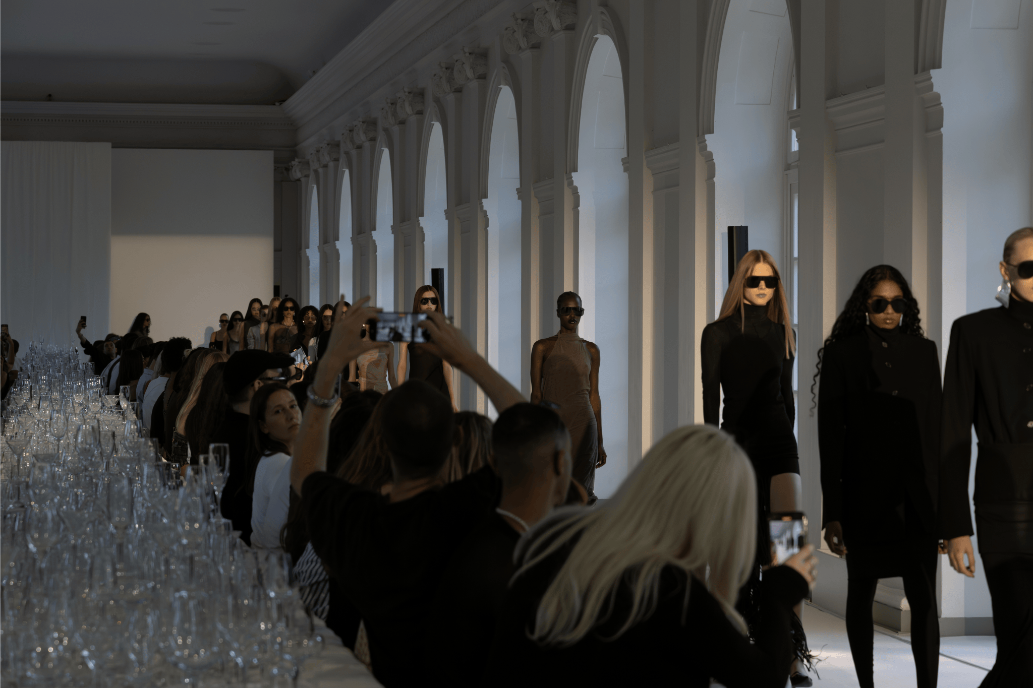  Models walking down the runway in dark-toned outfits during the Ioannes SS26 show at Berlin Fashion Week, with an audience capturing the moment on their phones.