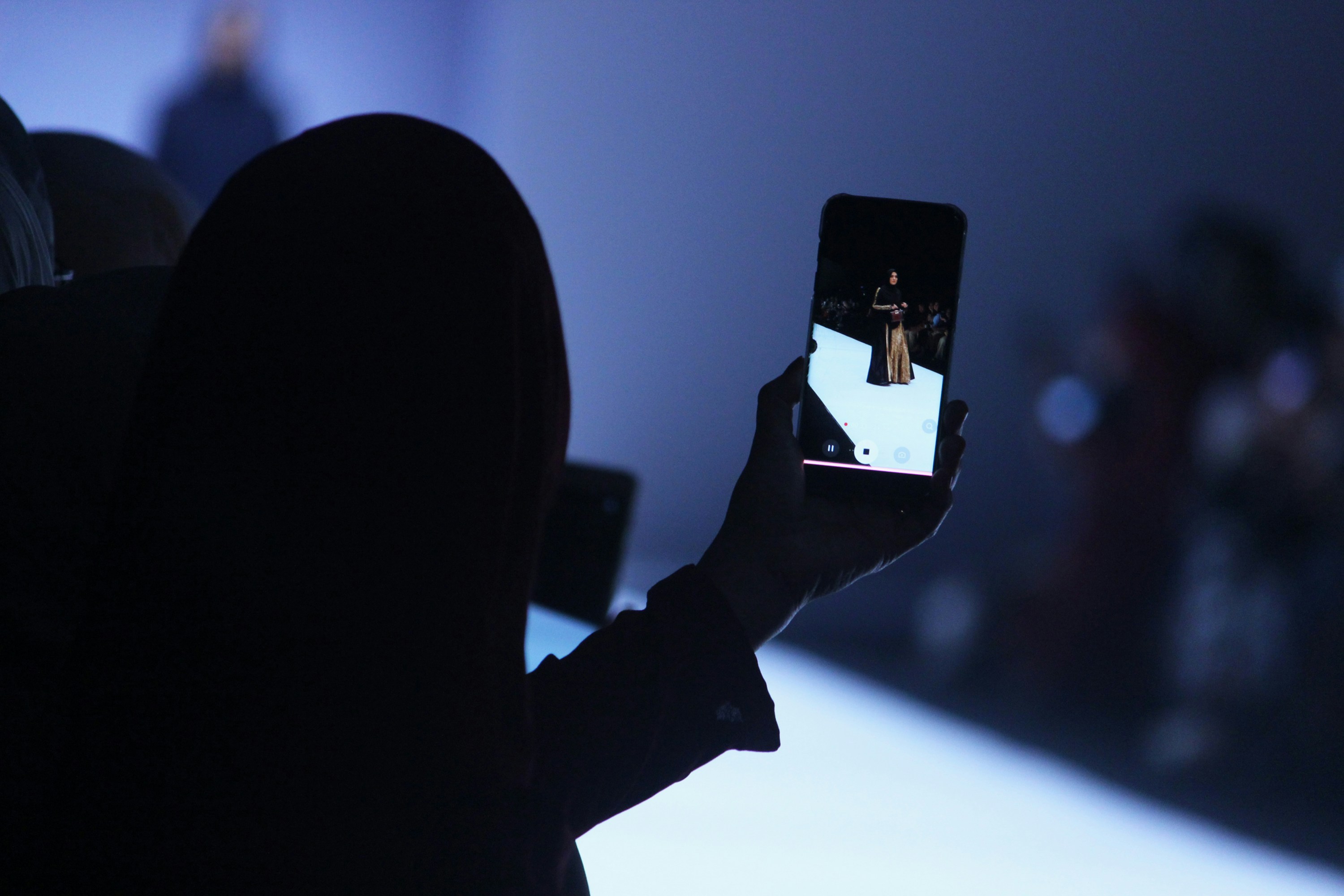  A front-row guest captures a model walking the runway on their smartphone during a fashion show, silhouetted against a blue-lit background.