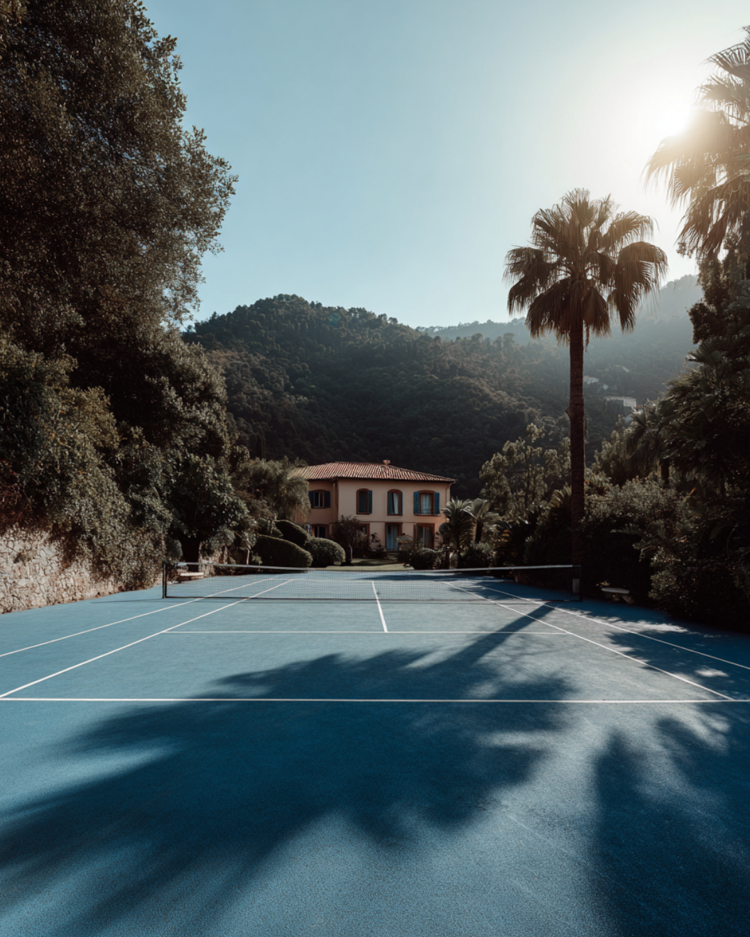 Elegant Mediterranean tennis court surrounded by lush greenery and palm trees with a villa in the background under warm morning sunlight