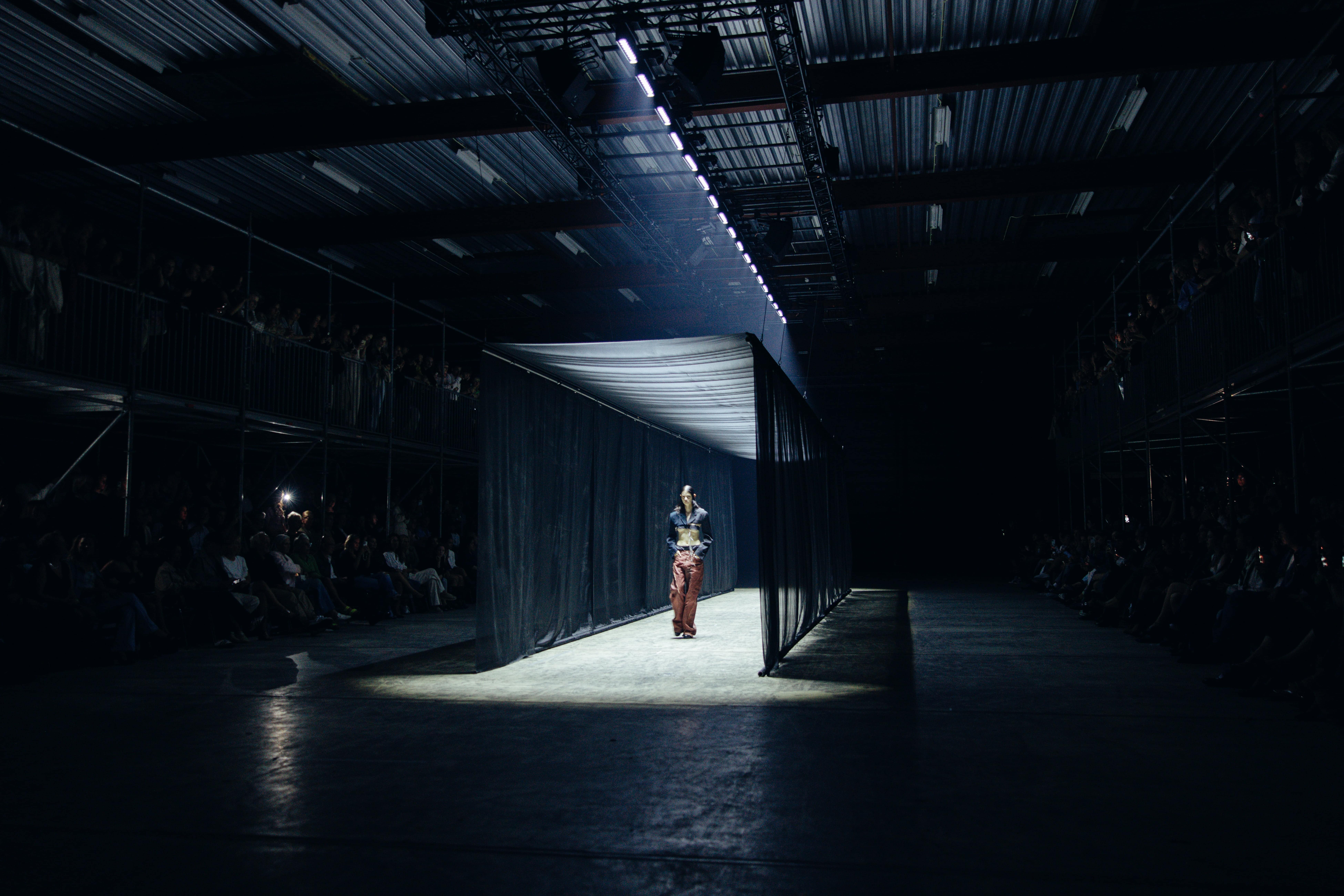 Model walks down a spotlighted runway surrounded by sheer black curtains at Amsterdam Fashion Week 2024, with an audience seated in a dark industrial venue.