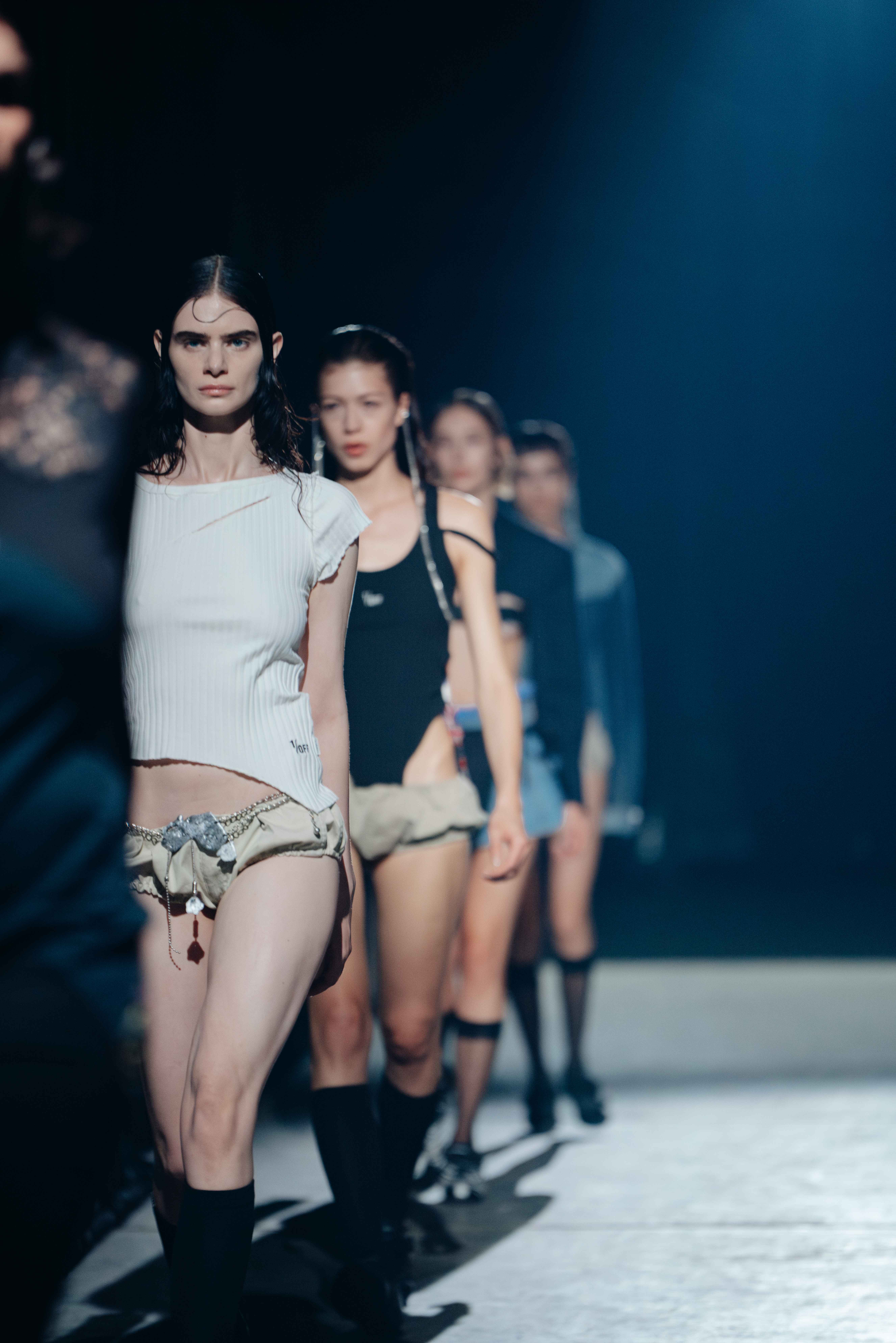 Models walk in a straight line during the 1/OFF show at Amsterdam Fashion Week 2024, wearing deconstructed and minimalistic outfits with raw-edge aesthetics under dramatic lighting.