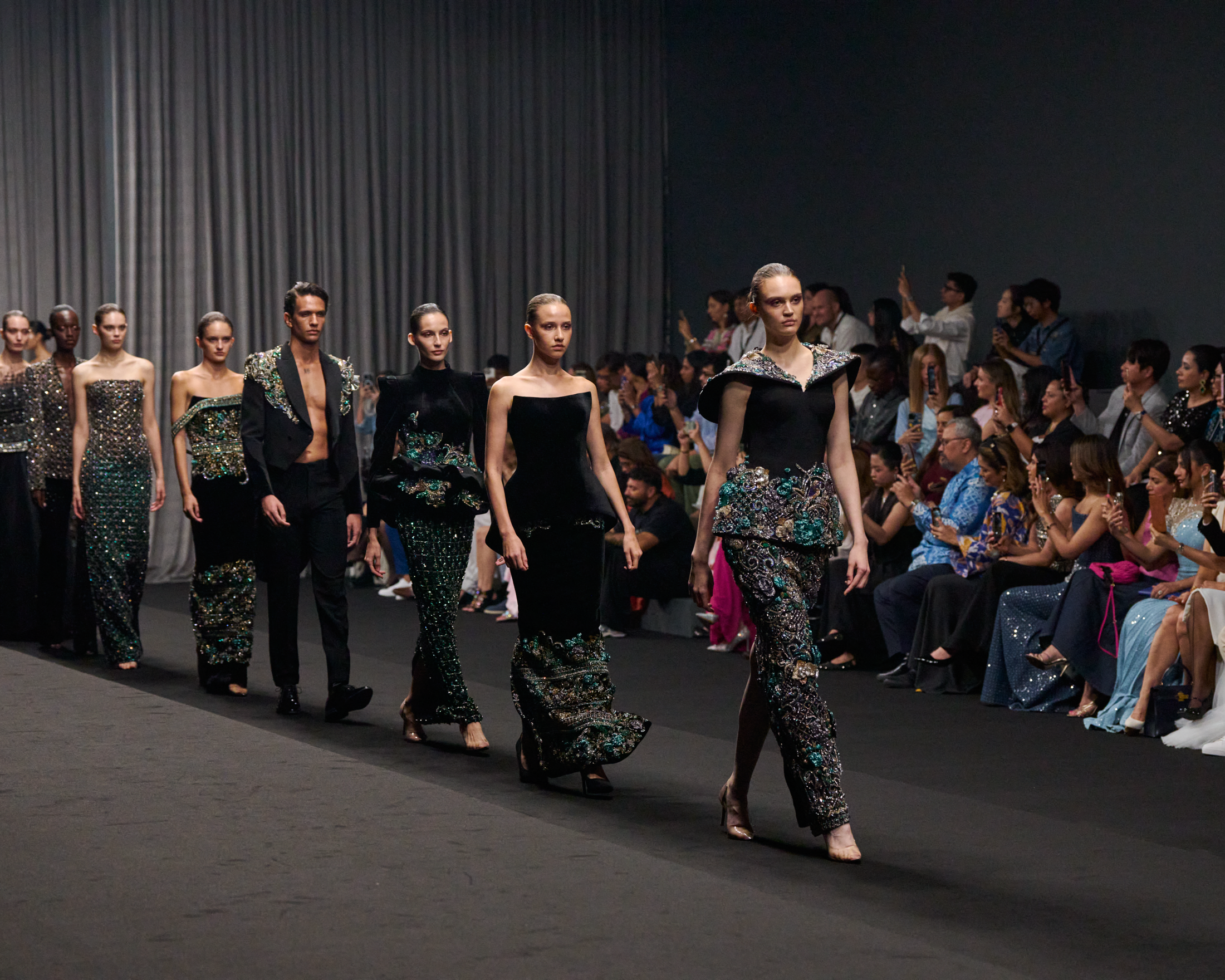 A group of models walking the runway during Ruzaini’s Haute Couture show at Dubai Fashion Week Spring/Summer 2026 in September 2025. The models wear black and emerald green embellished gowns and suits, with a captivated audience in the background.