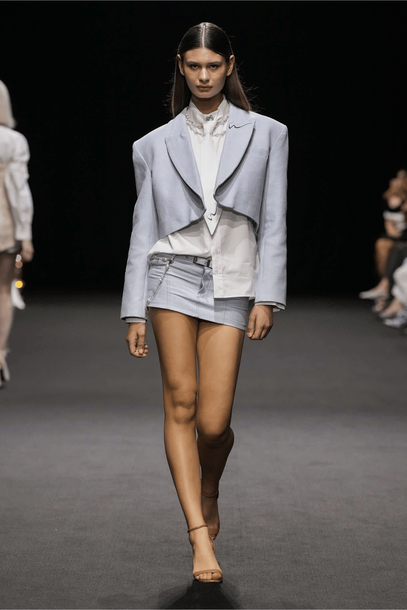 A model wearing a short denim skirt, white blouse, and structured light blue blazer walks the runway at the Weinsanto AW25/26 show during Dubai Fashion Week Autumn/Winter 2025–2026.