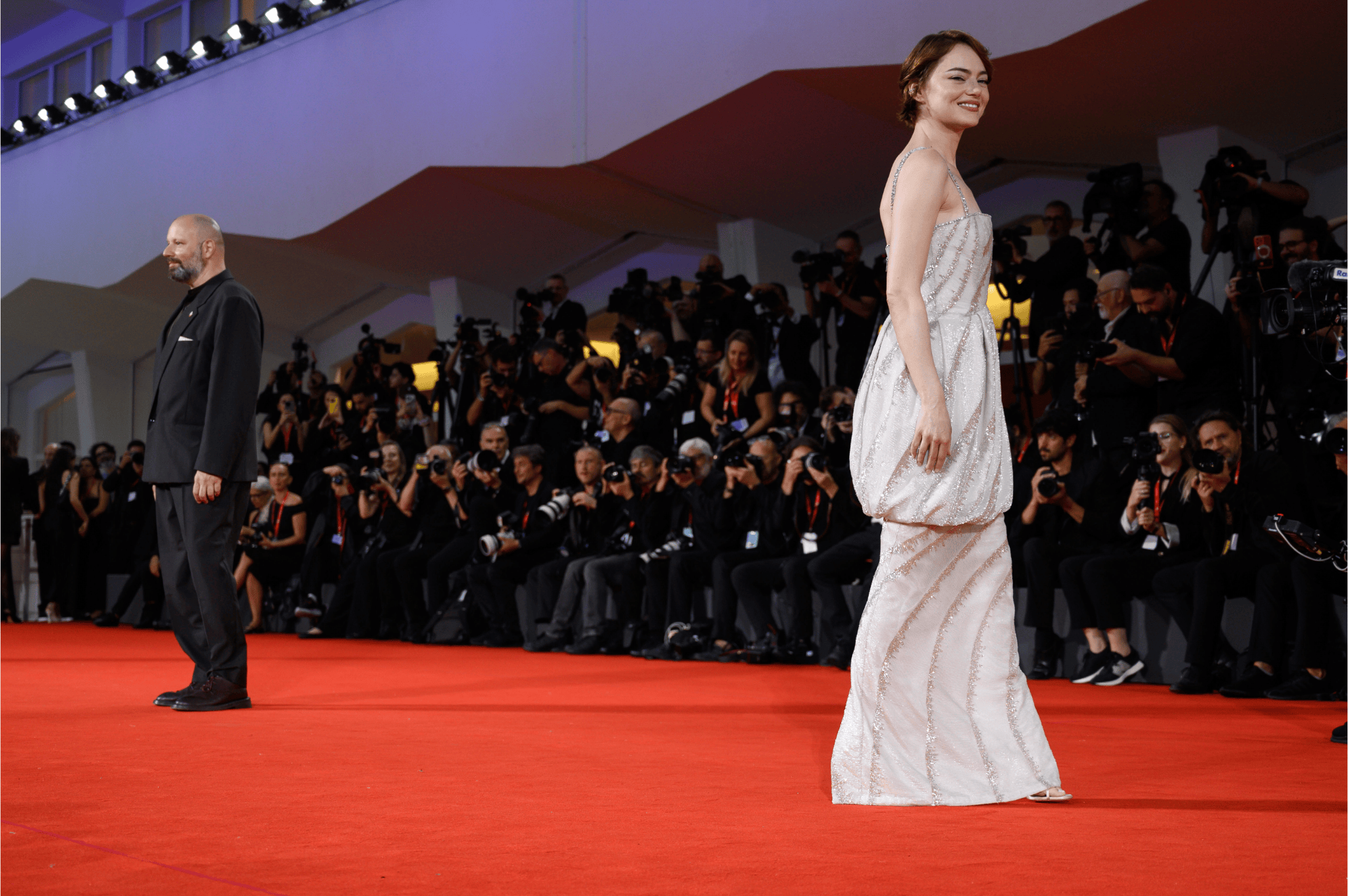 Actress Emma Stone wearing an elegant gown, smiles on the red carpet during the premiere of Bugonia at the 82nd Venice International Film Festival, while photographers capture the moment.