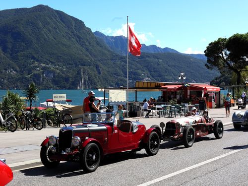 Drivers cruise vintage sports cars along the Campione lakeside road, passing cafes and the Swiss flag with the stunning Lake Lugano and mountains as a backdrop.