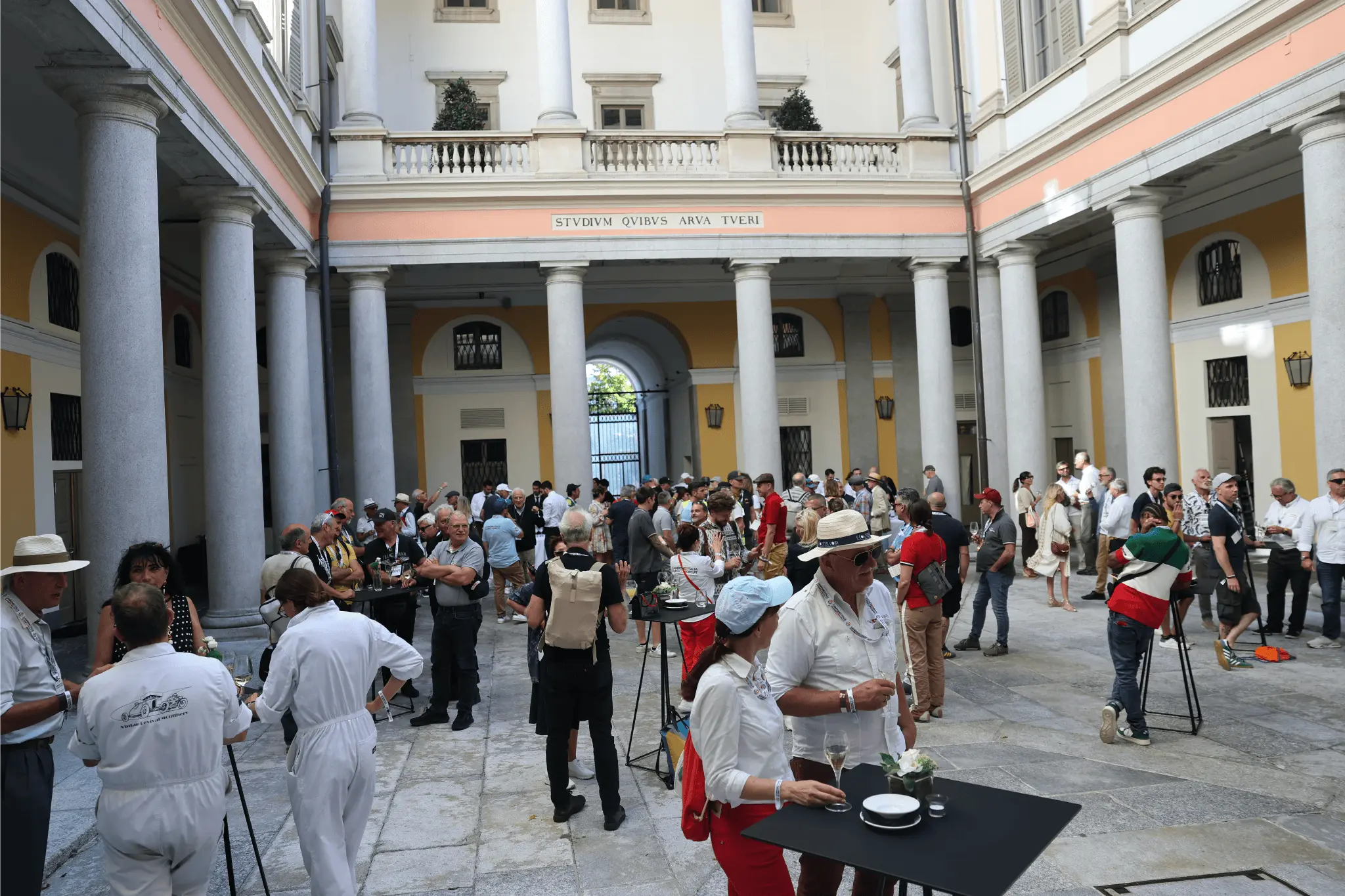A large group of participants and guests enjoy an aperitif under the colonnade of Lugano City Hall during the Campione d’Italia Classic Car Circuit 2025. The courtyard is filled with vintage car enthusiasts chatting and holding glasses, set against historic architecture.