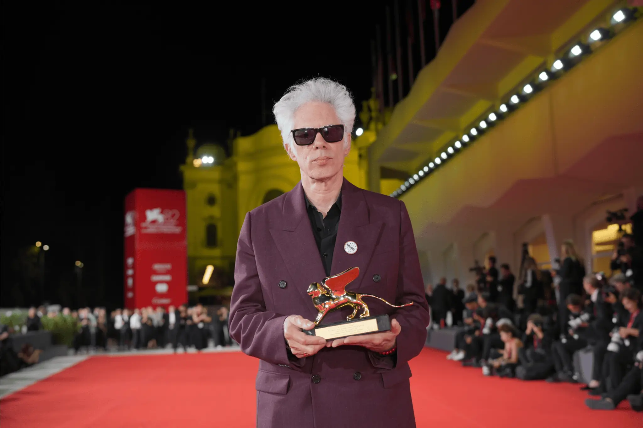 Director Jim Jarmusch holds the Golden Lion award on the red carpet at the 82nd Venice International Film Festival 2025, wearing sunglasses and a burgundy suit.