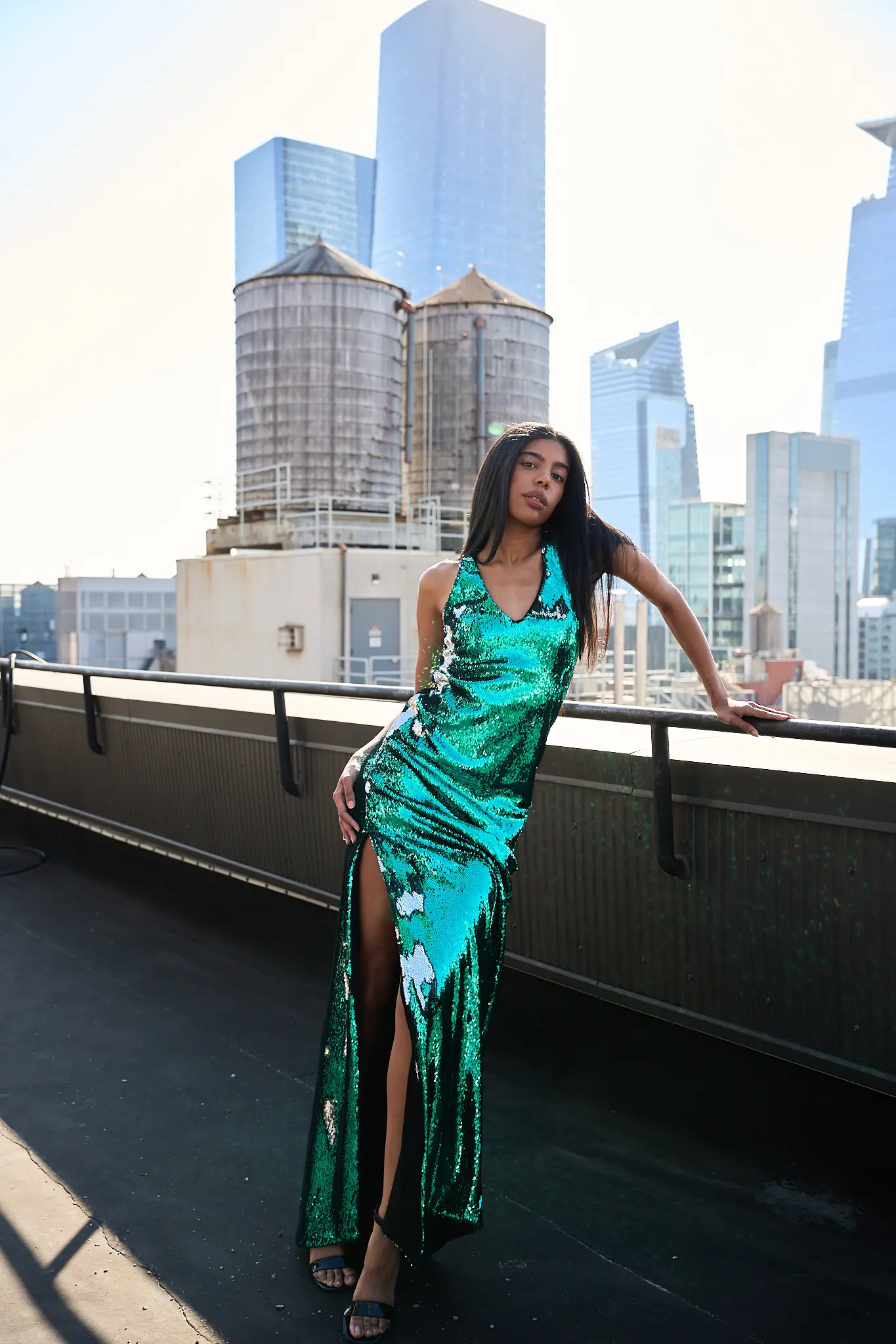 Model wearing a shimmering emerald green sequin gown with thigh-high slit by Pellone, photographed outdoors with a city skyline backdrop.
