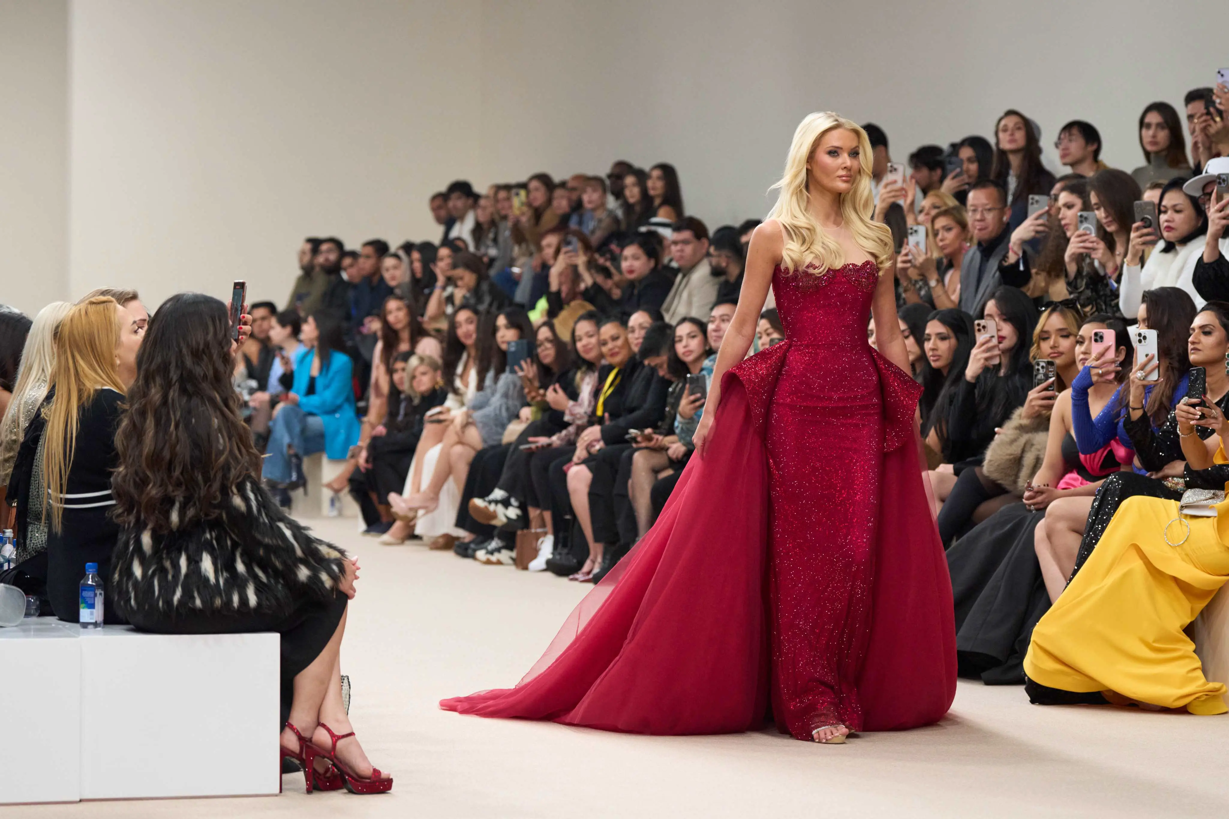 A blonde model in a sparkling deep red couture gown with a dramatic train walks the runway during Michael Cinco's Autumn/Winter 2025–26 show at Dubai Fashion Week, as a glamorous front-row audience captures the moment