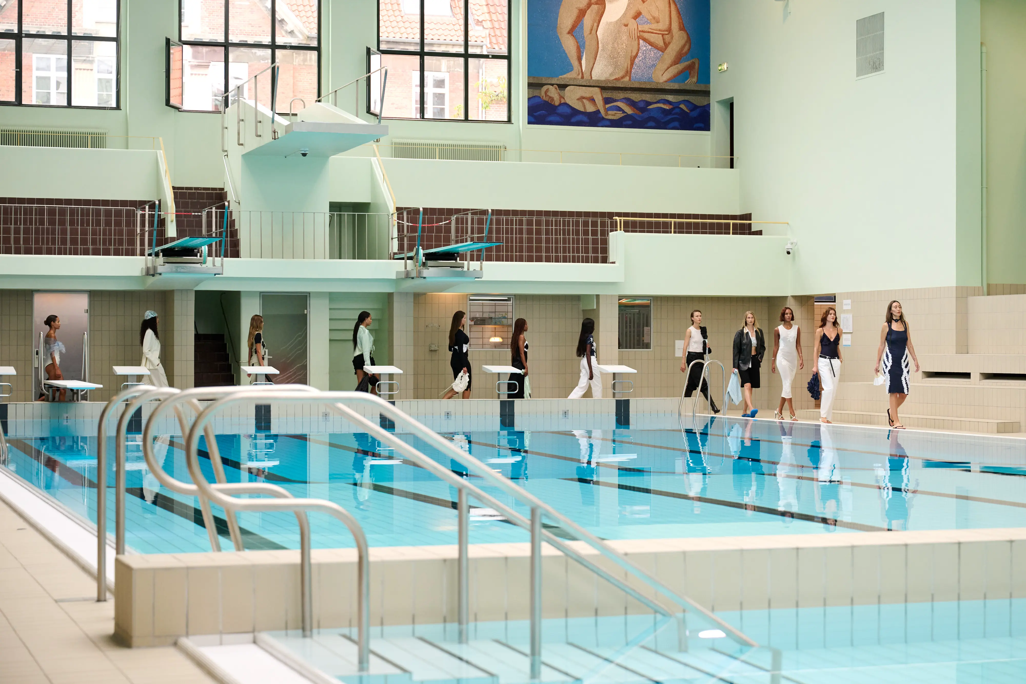 Models walk in single file beside an indoor swimming pool in sleek monochrome and navy outfits during OpéraSPORT’s Spring/Summer 2026 runway presentation at Copenhagen Fashion Week.