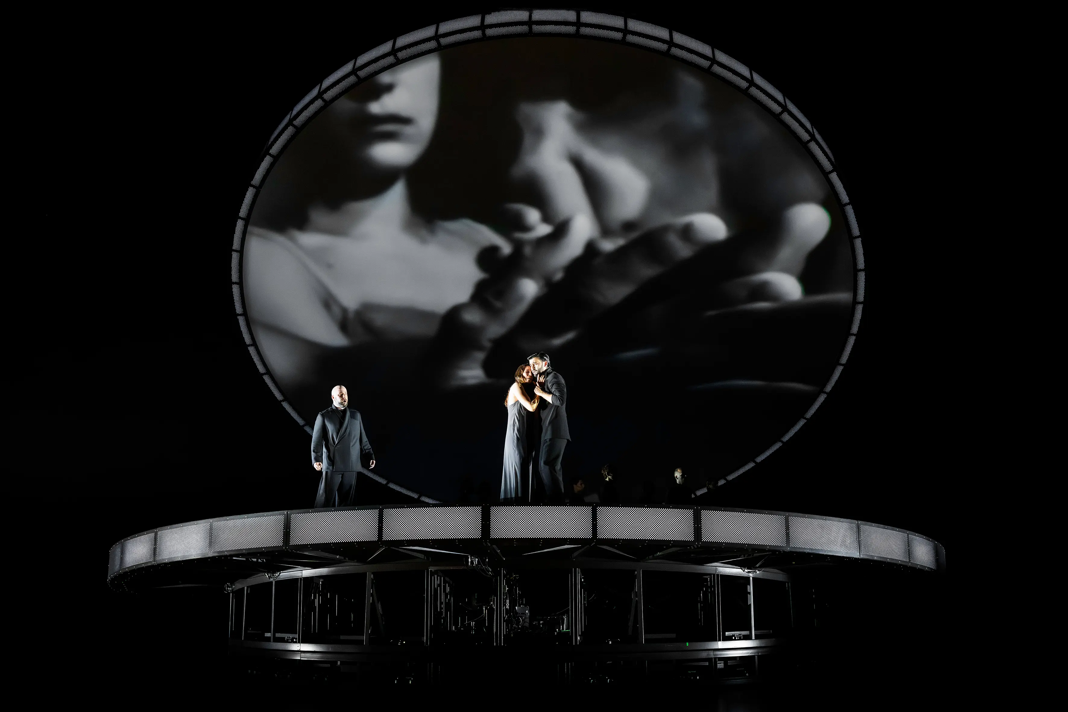 Two opera performers embracing on a futuristic circular stage with a cinematic projection above.