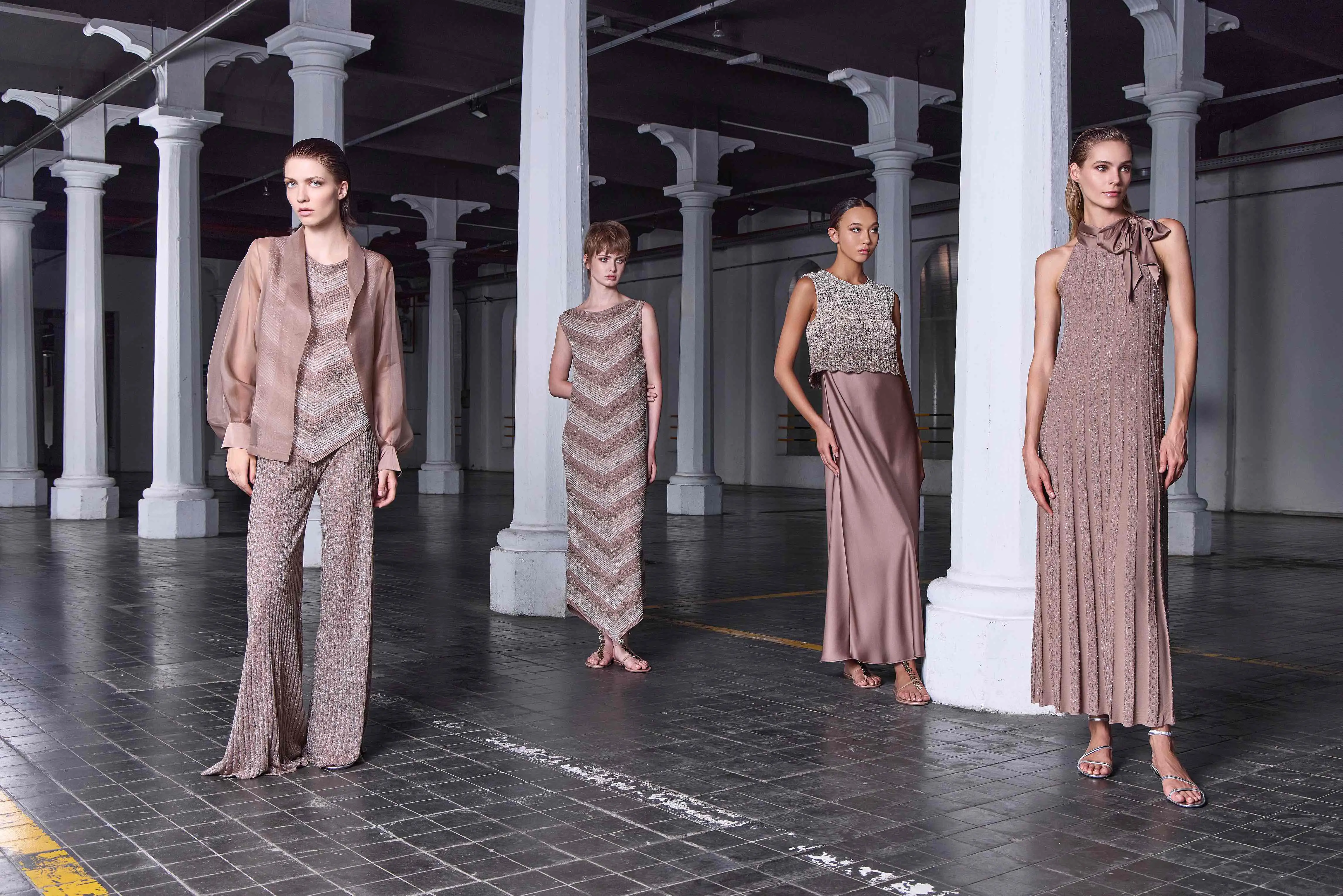 Four female models showcase minimalist, neutral-toned eveningwear in a modern industrial setting during La Moda Italiana Vol. II at Dubai Fashion Week September 2025. Outfits include pleated pants, shimmering gowns, and sleeveless knits in soft taupe hues.
